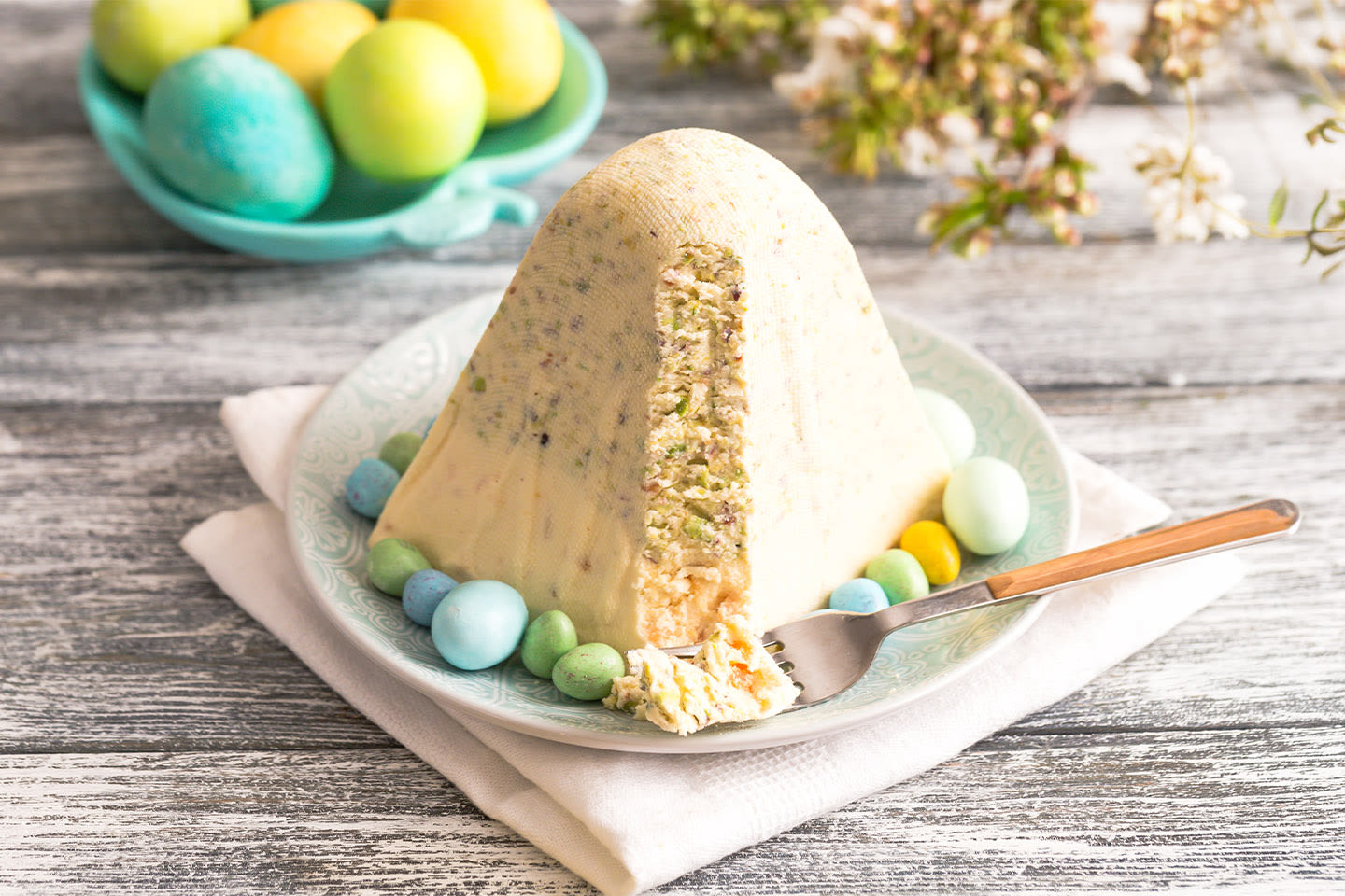 Pääsiäisen rahkapasha lautasella värikkäiden suklaamunien ympäröimänä, taustalla turkoosilla vadilla värjättyjä kananmunia.