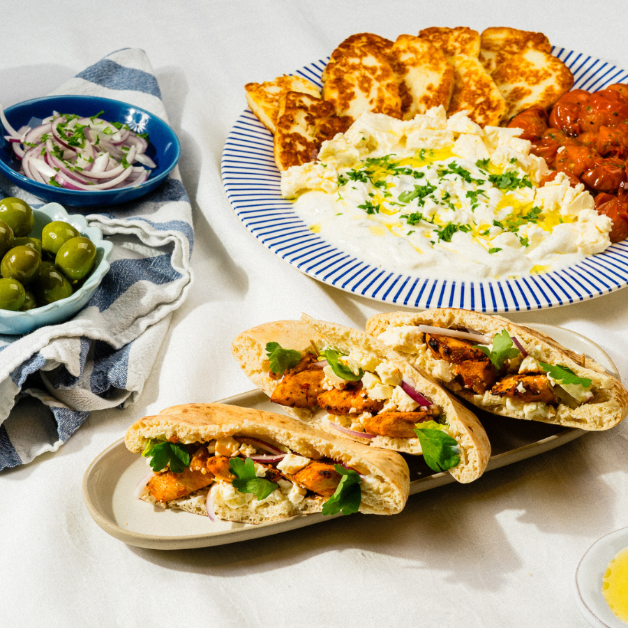 Herkullinen kreikkalainen ateria: pitaleipää täytettynä kanalla, siniraitainen lautanen fetajuustolla ja pikkukulhot sipulilla ja oliiveilla.