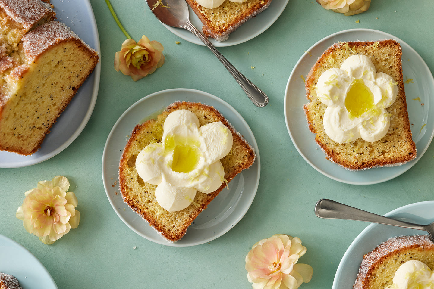 Mehevää kakkua vaaleansinisellä pöydällä, siivut koristeltu kermakukilla ja vieressä hentoja vaaleanpunaisia kukkia.