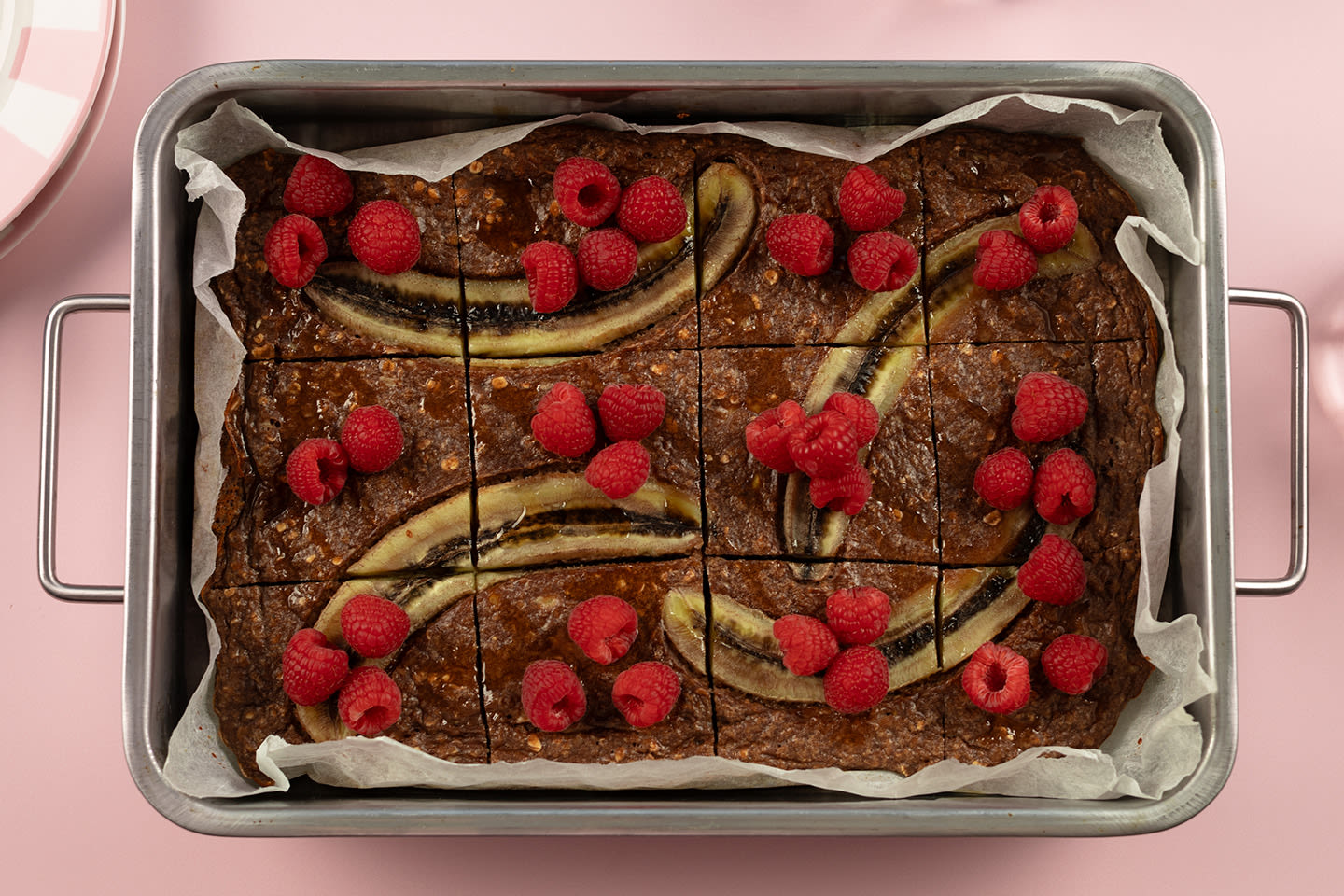 Mehevä suklaakakku pellillä koristeltuna tuoreilla vadelmilla ja banaanisiivuilla, valmiiksi paloiteltuna.