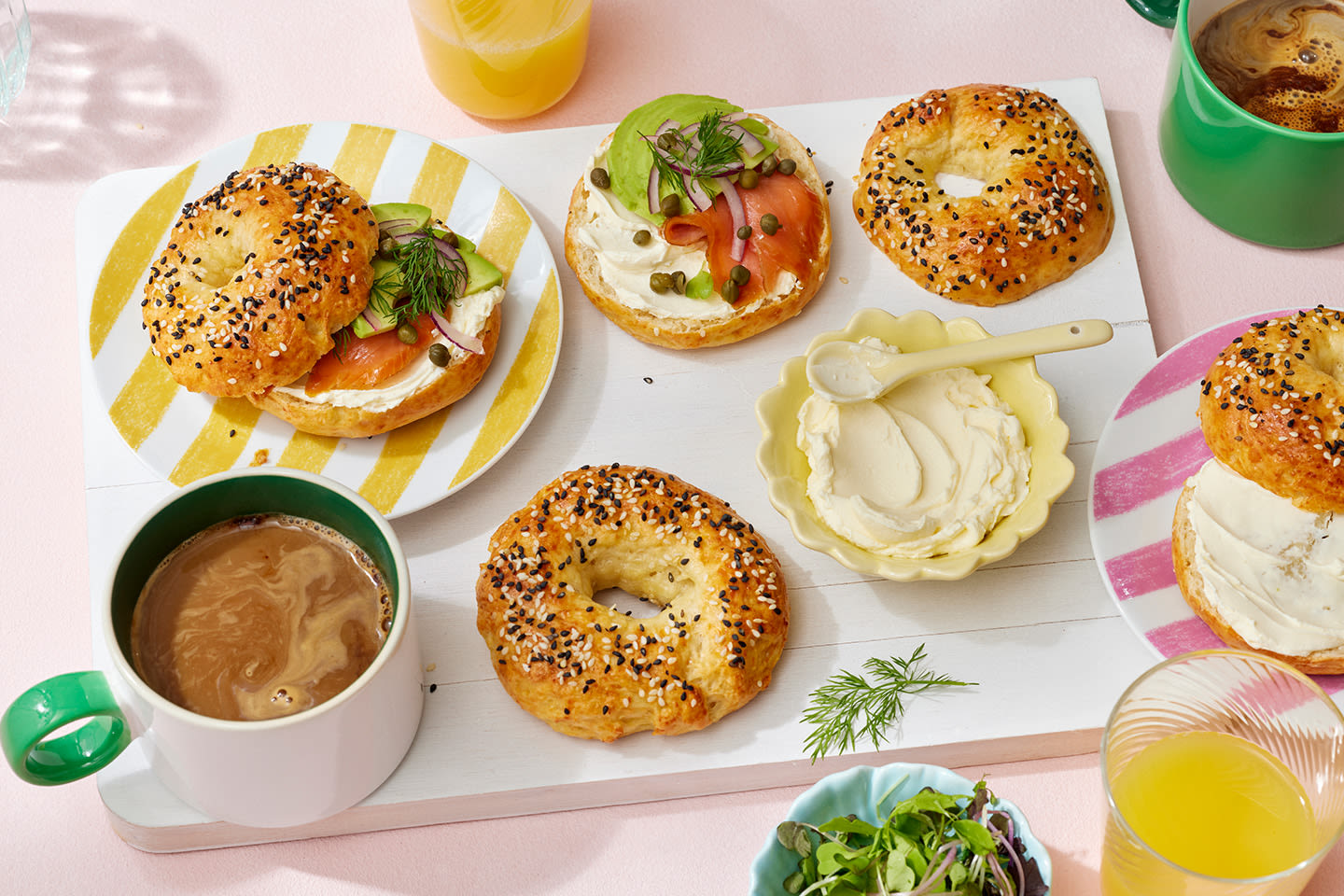 Aamiaiskattaus, jossa siemenillä koristeltuja bageleita, tuorejuustoa ja lohta, kahvia ja mehua vaaleanpunaisella pöydällä.