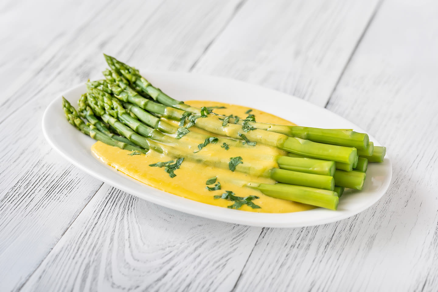 Mehevää parsaa valkoisella lautasella, päällä keltaista hollandaisekastiketta ja tuoretta yrttiä, vaalealla puupöydällä.