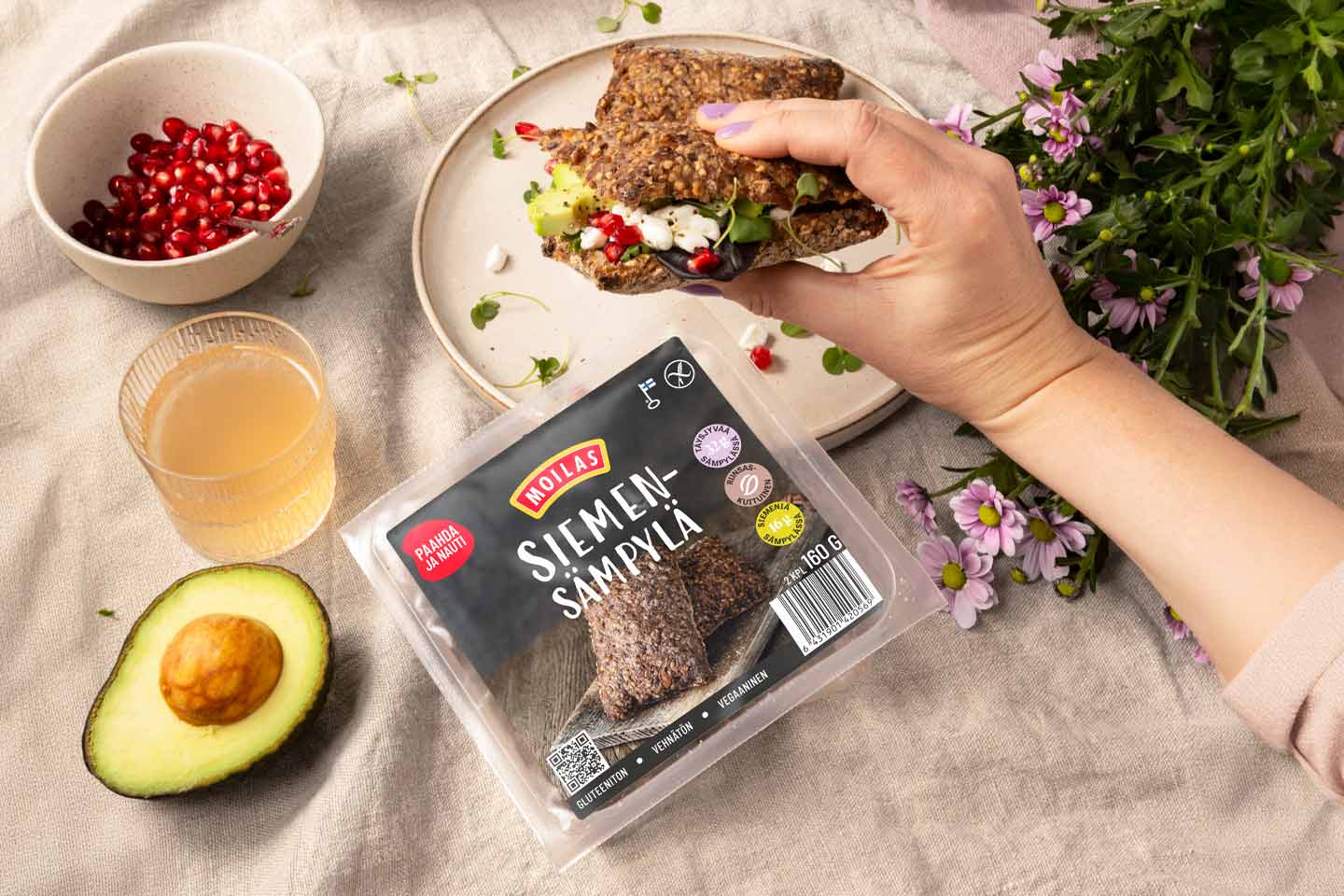 Käsi pitää täytettyä Moilas-siemensämpylää lautasen päällä, vieressä Moilas-siemensämpyläpakkaus, avokado, granaattiomenansiemeniä, mehulasi ja kukkia.