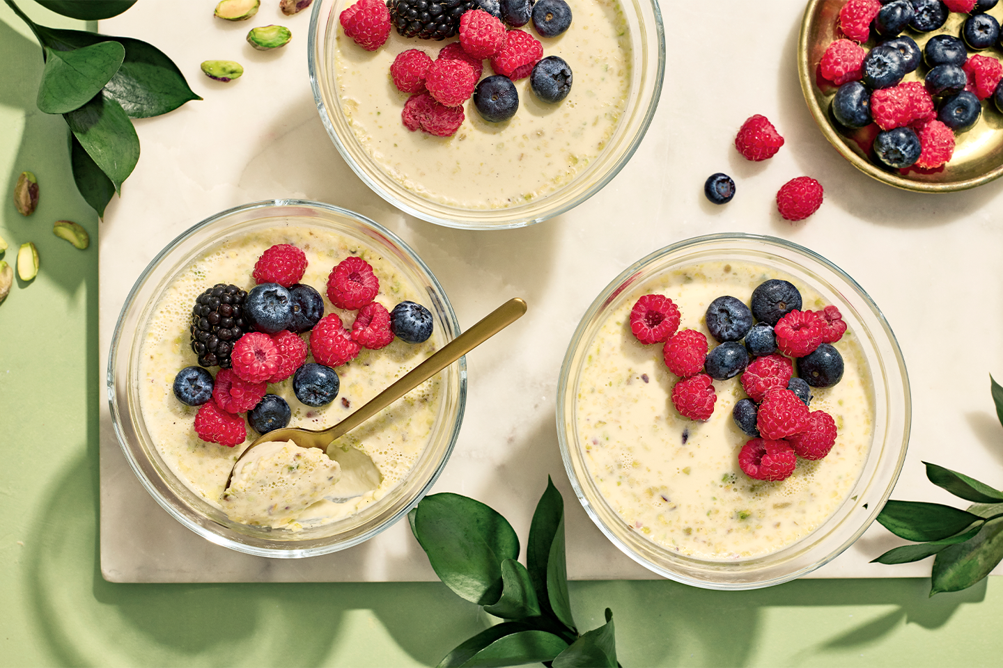 Herkullisia pistaasipannacotta-annoksia tuoreilla vadelmilla, mustikoilla ja karhunvatukoilla koristeltuna vihreillä lehdillä somistettuna.