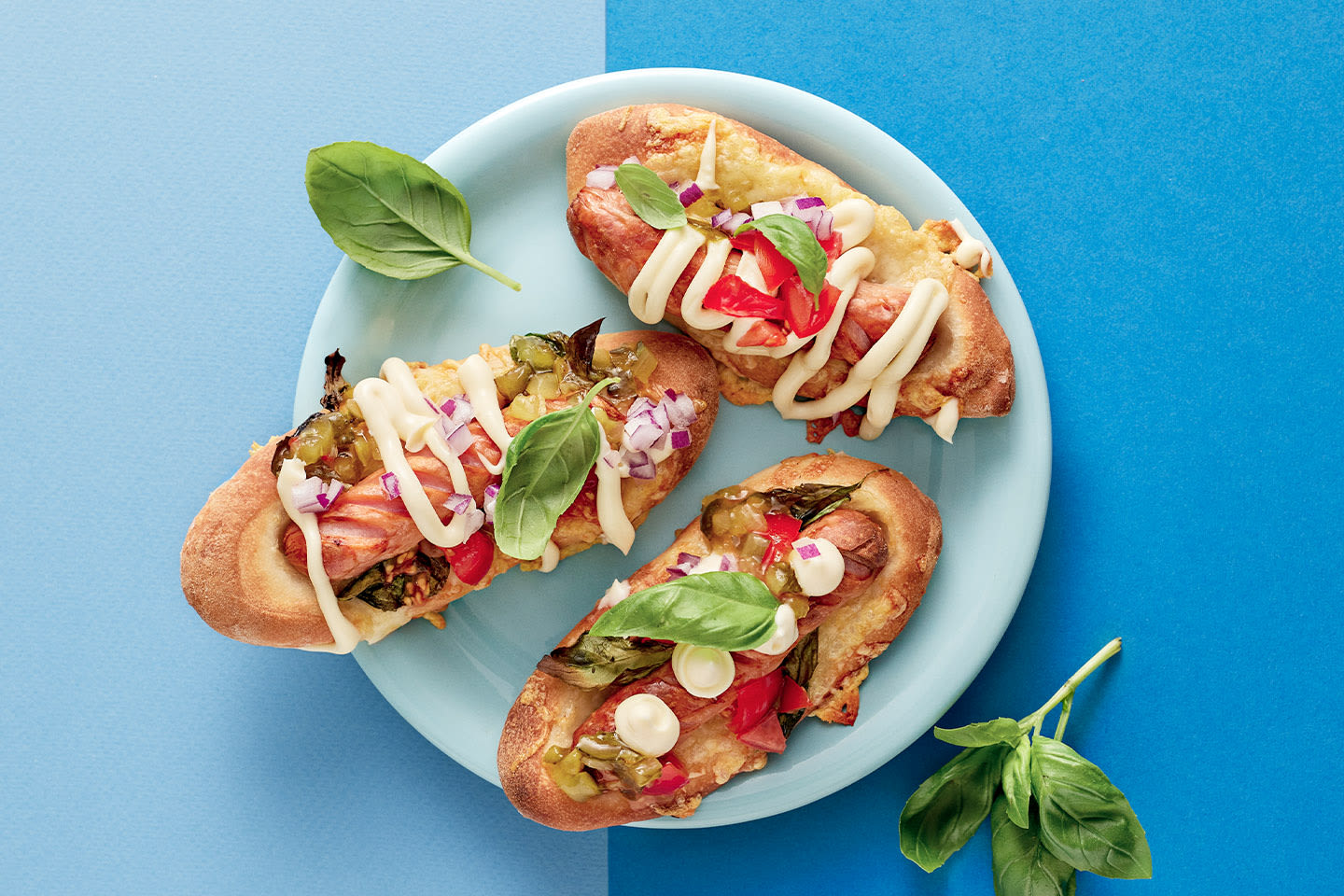 Kolme herkullista pizzahodaria vaaleansinisellä lautasella, koristeltuna majoneesilla, punasipulilla ja tuoreella basilikalla.