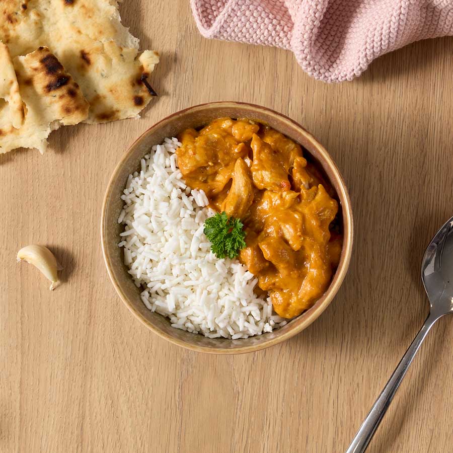 Kulhossa on riisiä ja oranssia kana-currya, vieressä naan-leipää ja lusikka puupöydällä.