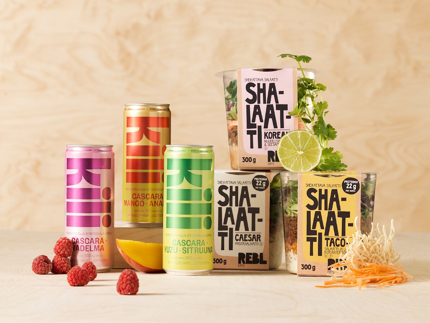 Raikkaita Shalaatti-salaatteja ja värikkäitä Kili-juomatölkkejä vadelmien, limetin ja korianterin kera vaaleaa taustaa vasten.