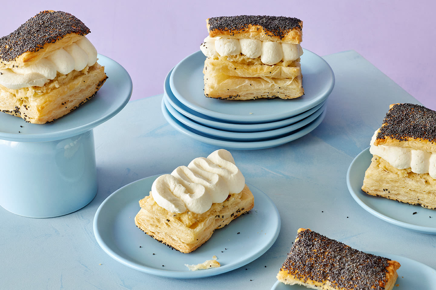 Herkullisia leivonnaisia vaaleansinisillä lautasilla - kermaisia unikkoleipäleivoksia kermavaahdon kera kevyessä asetelmassa.