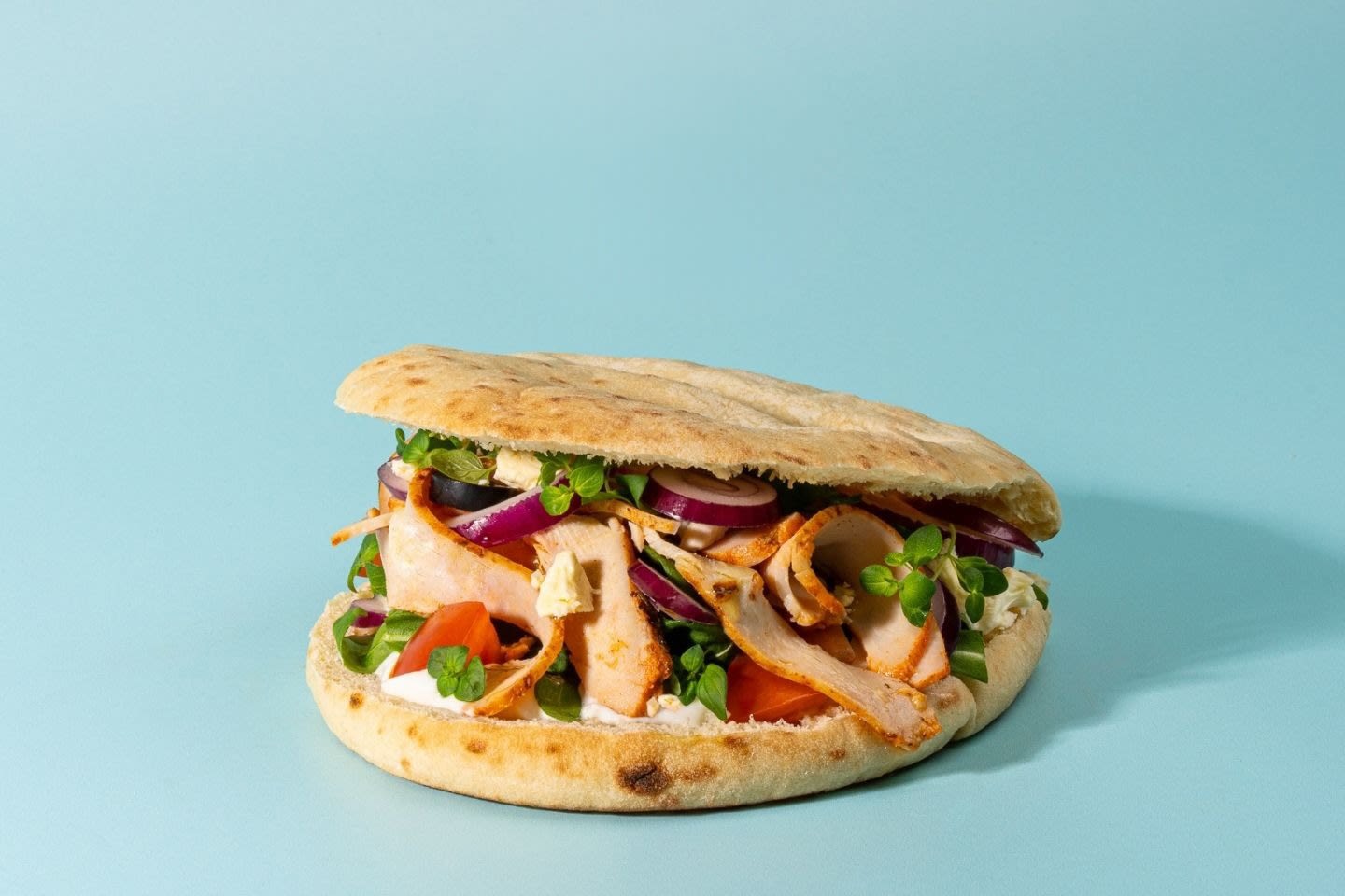 Mehevä pitaleipä täytetty kanalla, punasipulilla, tomaatilla ja tuoreilla yrteillä vaaleansinistä taustaa vasten.