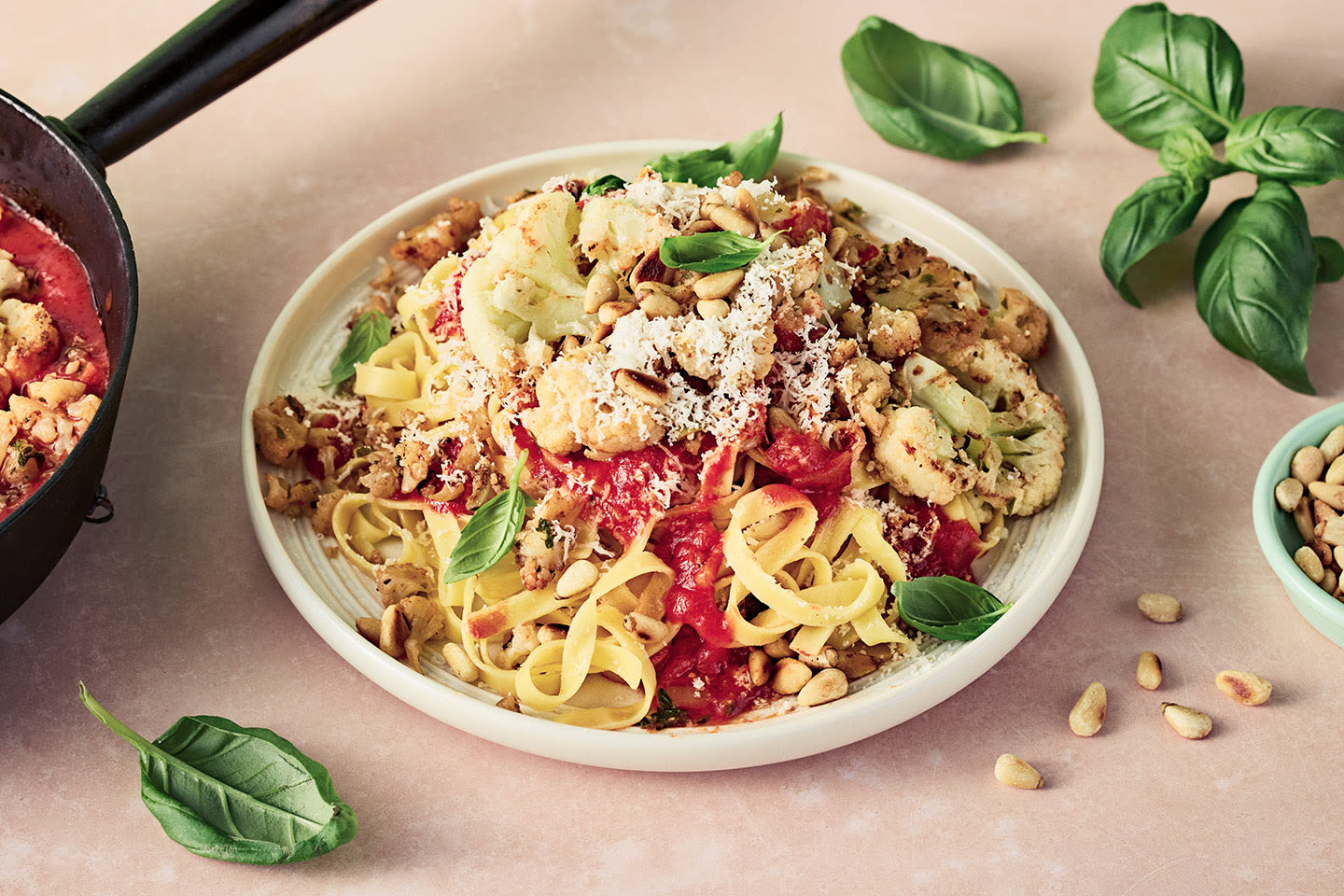 Lautasellinen tagliatelle-pastaa tomaattikastikkeella, paahdetulla kukkakaaleilla, pinjansiemenillä ja raastetulla juustolla, koristeltuna basilikalla.