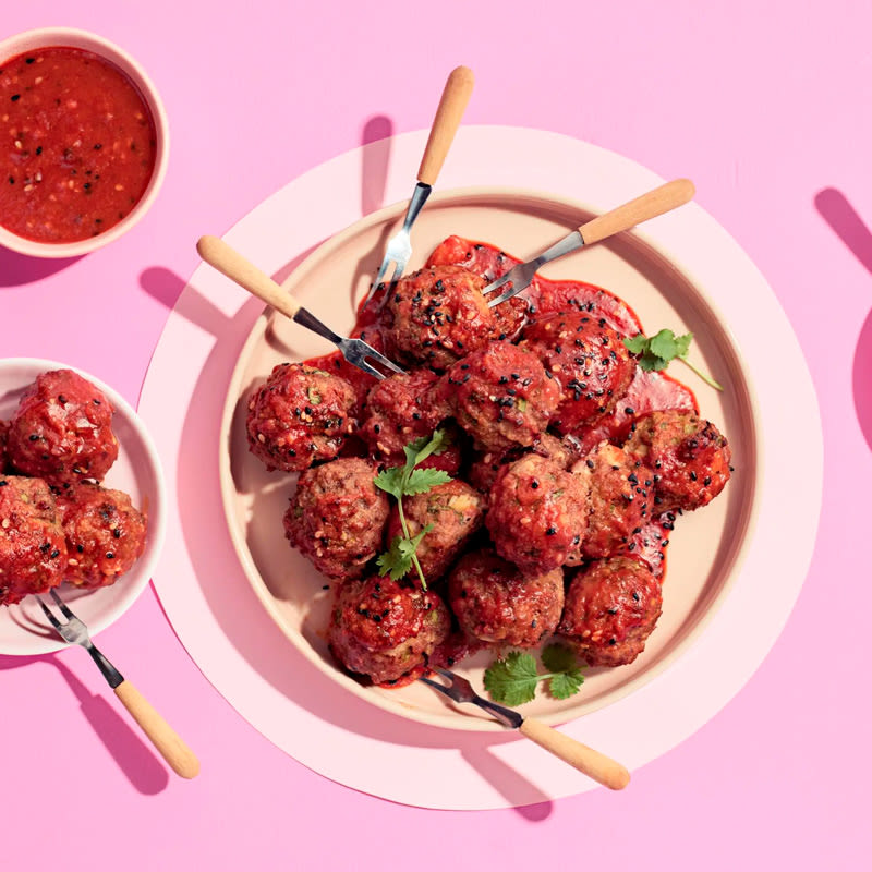Mehukkaita lihapullia vaaleanpunaisella taustalla, tarjoiltuna valkoisella lautasella pikkuhaarukoiden ja kastikkeen kera.