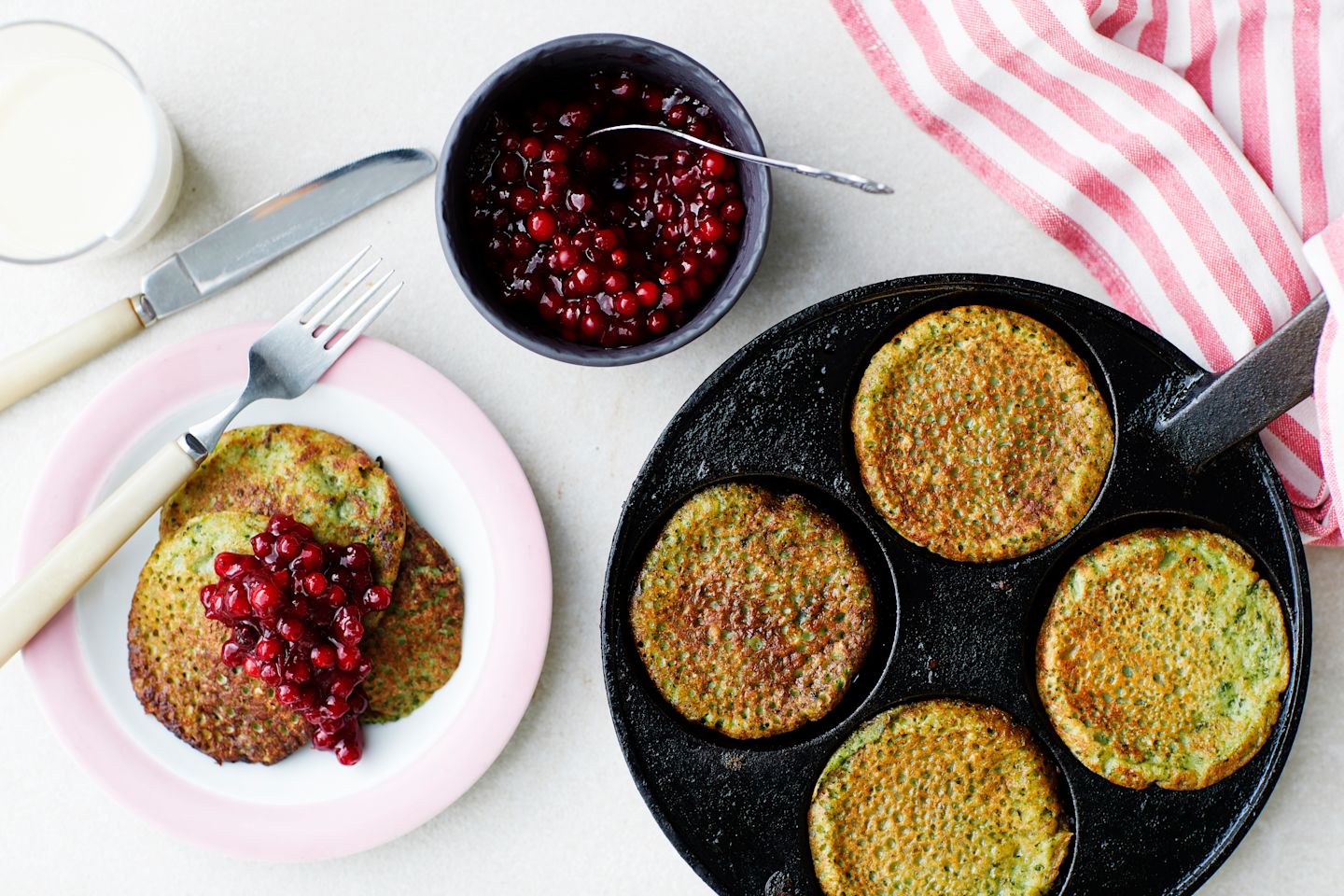 Rapeita lettuja valurautapannulla, vieressä vaaleanpunaisella lautasella lettu puolukkahillolla ja kulhossa lisää hilloa.