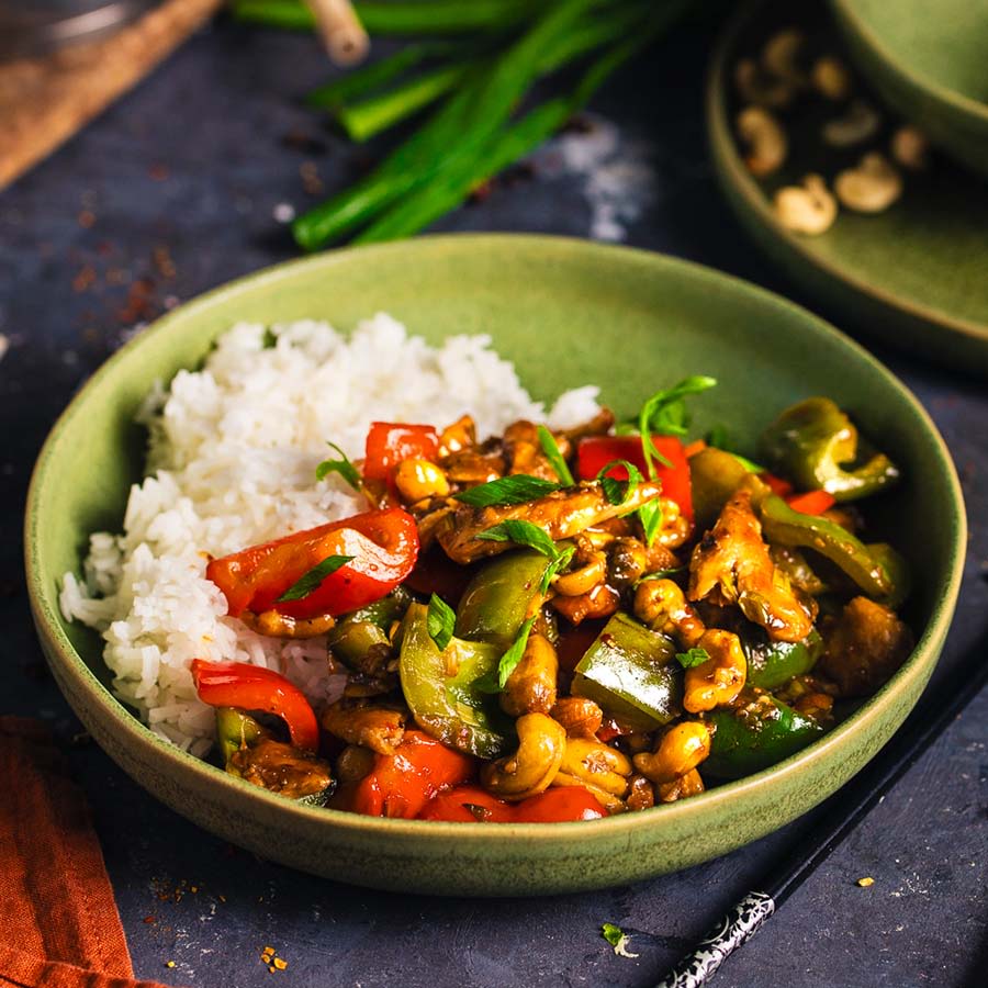 Vihreässä kulhossa on riisiä ja värikästä wok-paistosta kasviproteiinisuikaleilla, kasviksilla, cashew-pähkinöillä ja kastikkeella.