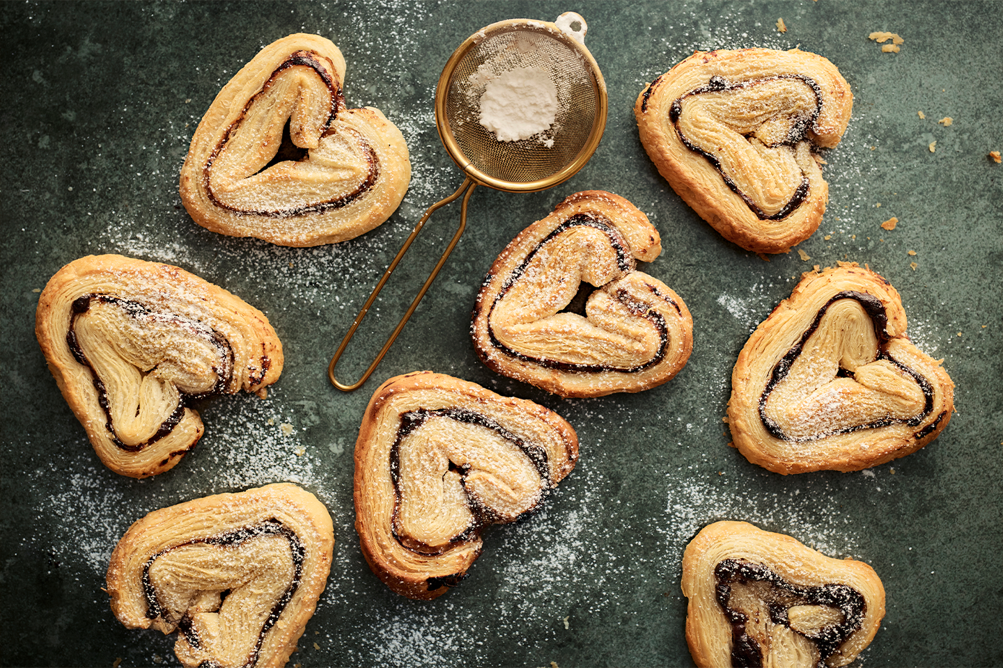 Sydämenmuotoisia leivonnaisia tomusokerilla ripoteltuna tummalla alustalla, keskellä pieni siivilä.