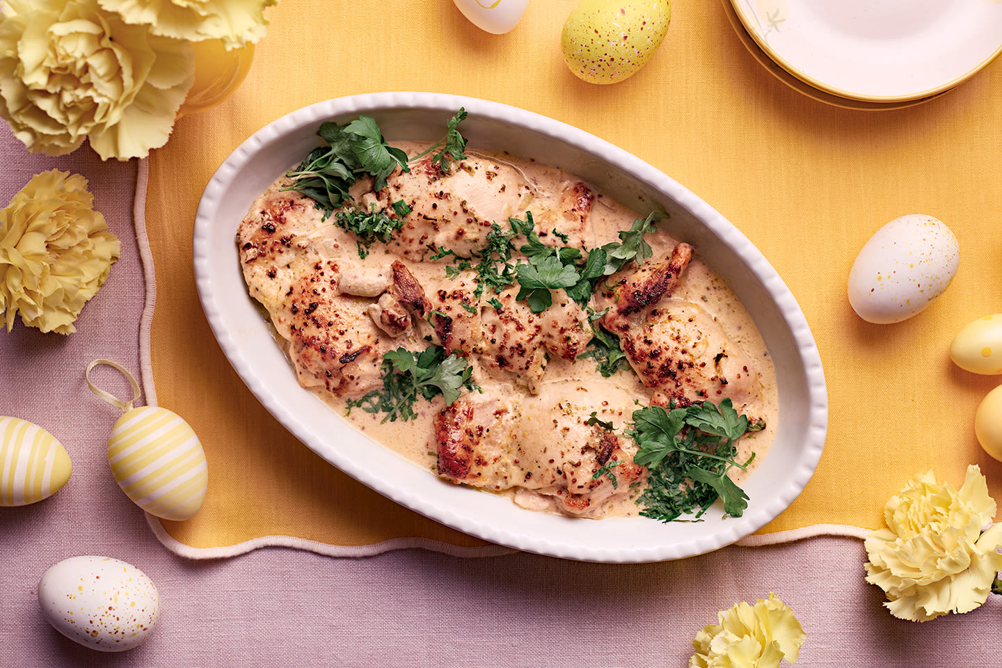 Mehevää kanaa kermaisessa kastikkeessa vaalean keltaisella pöydällä, ympärillä pääsiäismunia ja keltaisia kukkia.