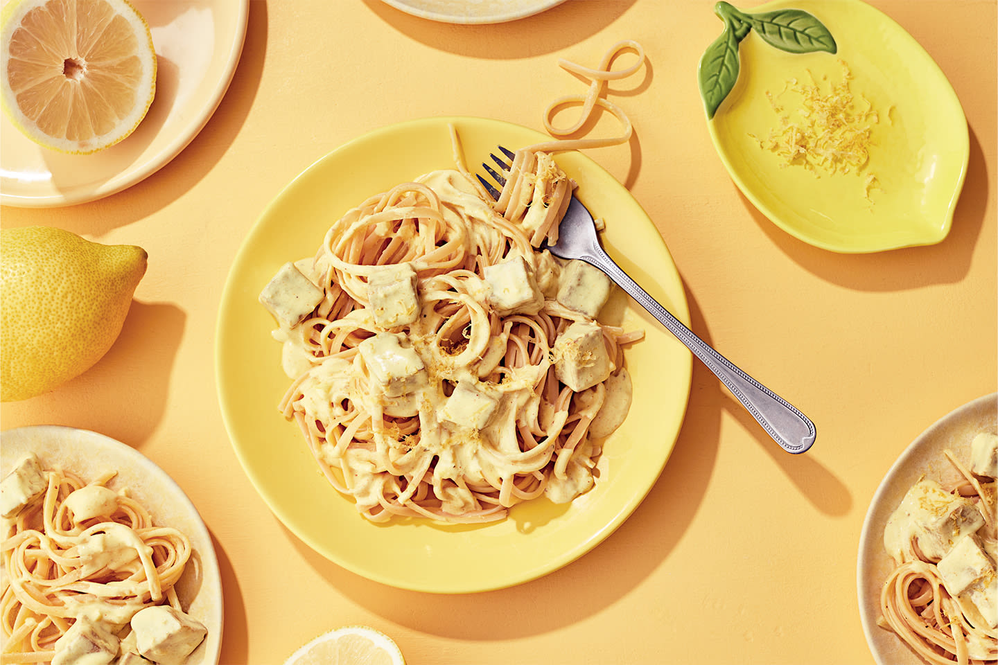 Keltaisella lautasella pastaa ja tofua sitruunaisessa kastikkeessa, ympärillä sitruunoita ja muita annoksia aurinkoisella taustalla.