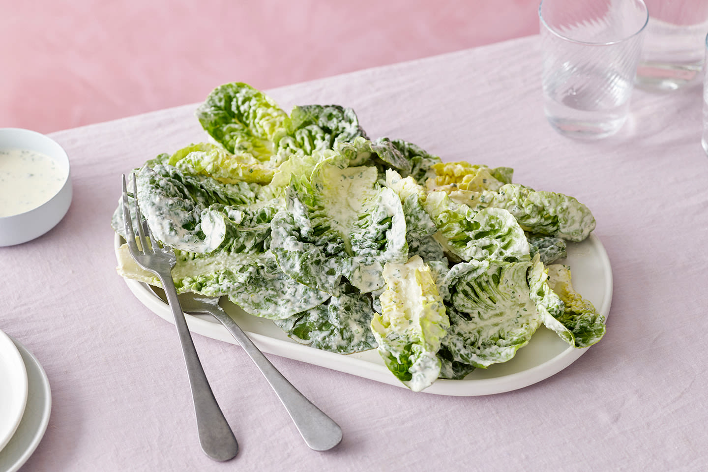 Kastikkeessa sekoitettua salaattia valkoisella lautasella, vieressä kastikekulho ja vesilaseja vaaleanpunaisella pöytäliinalla.