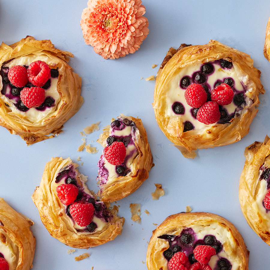 Vadelmilla, mustikoilla ja rahkalla täytettyjä kullanruskeita leivonnaisia sinisellä taustalla.