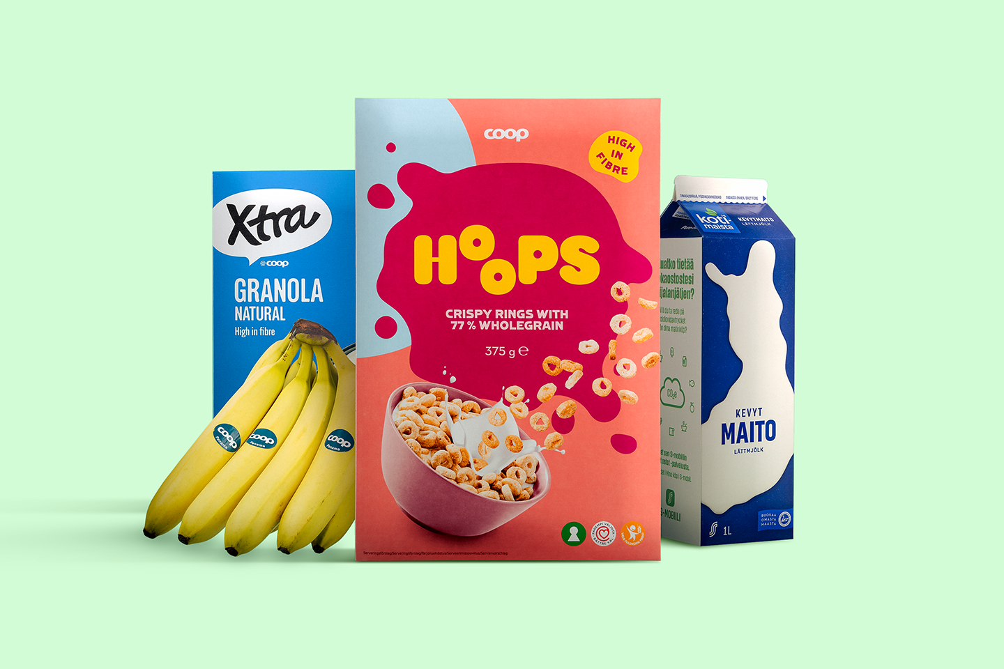 Aamiaistarvikkeita vihreällä taustalla: Hoops-murorenkaat, Xtra-granola, banaaneja ja kevytmaitoa .