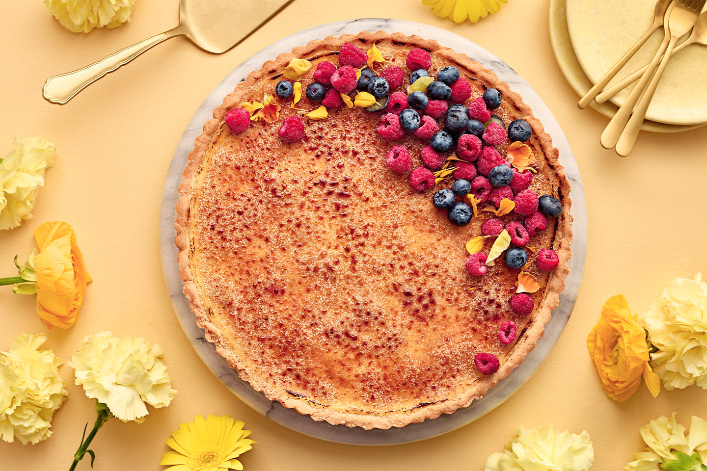 Herkullinen crème brûlée -piirakka koristeltuna mustikoilla ja vadelmilla keltaisella pöydällä, ympärillä kauniita kukkia ja kultaisia aterimia.