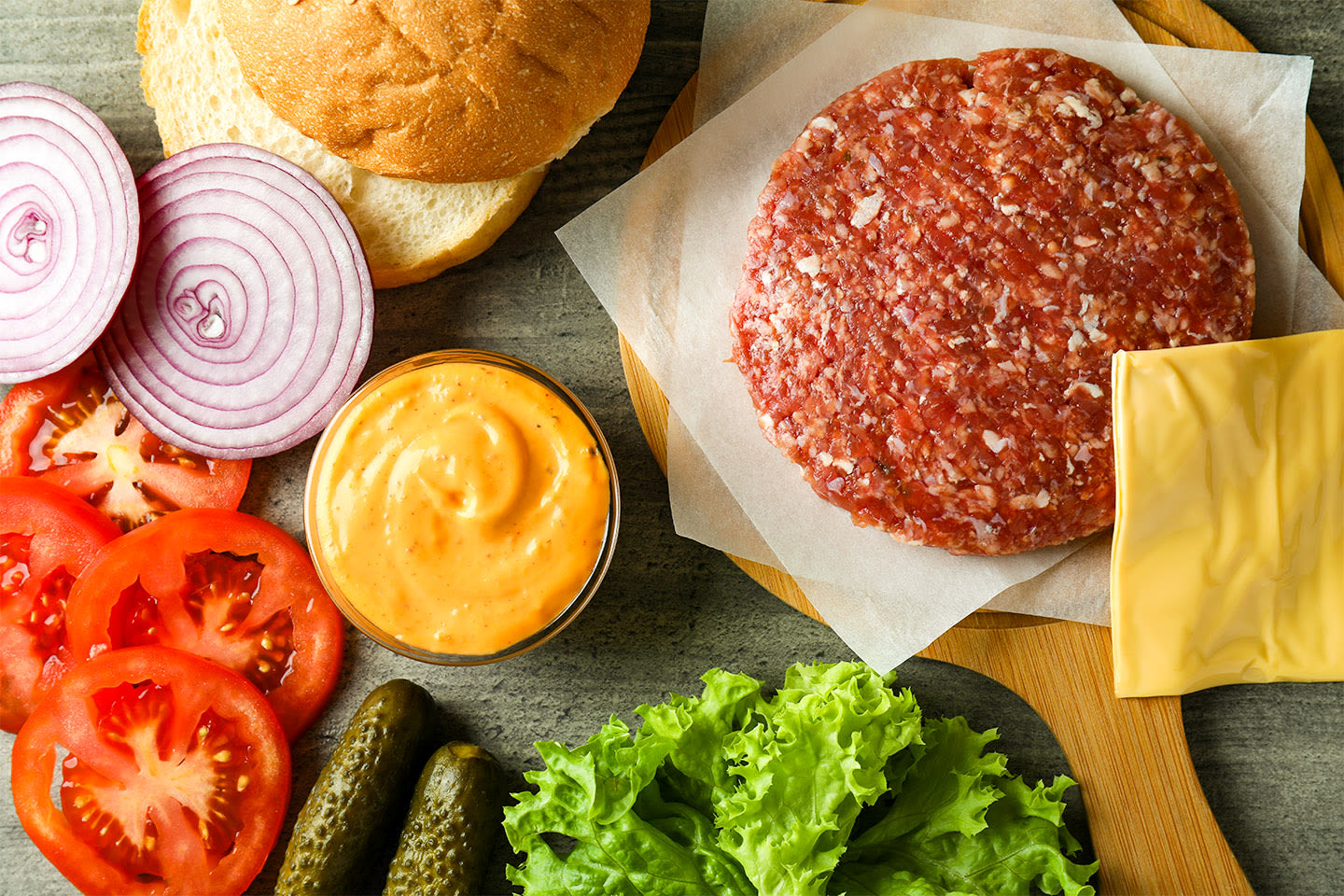 Raaka hampurilaispihvi ja tuoreet ainekset kuten sämpylä, juusto, tomaatti, sipuli, salaatti ja kastike valmiina kokoamista varten.