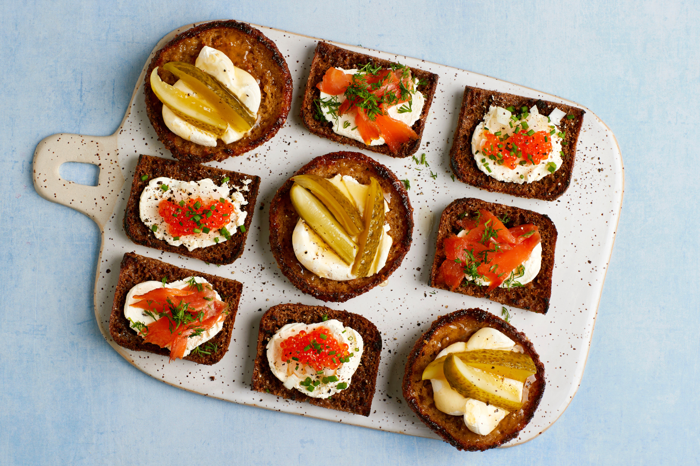 Herkullisia ruisleipäpaloja tarjoilulaudalla, päällä tuorejuustoa, lohta, mätiä ja suolakurkkua.