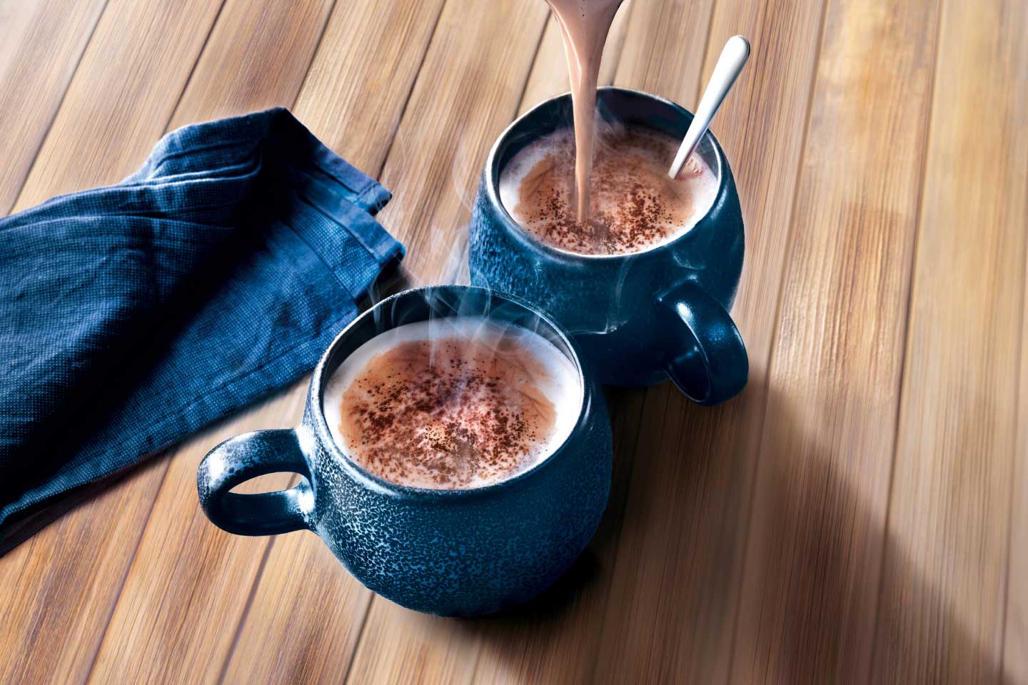 Kaakaota kaadetaan siniseen mukiin puisella pöydällä, vieressä toinen muki ja sininen liina.