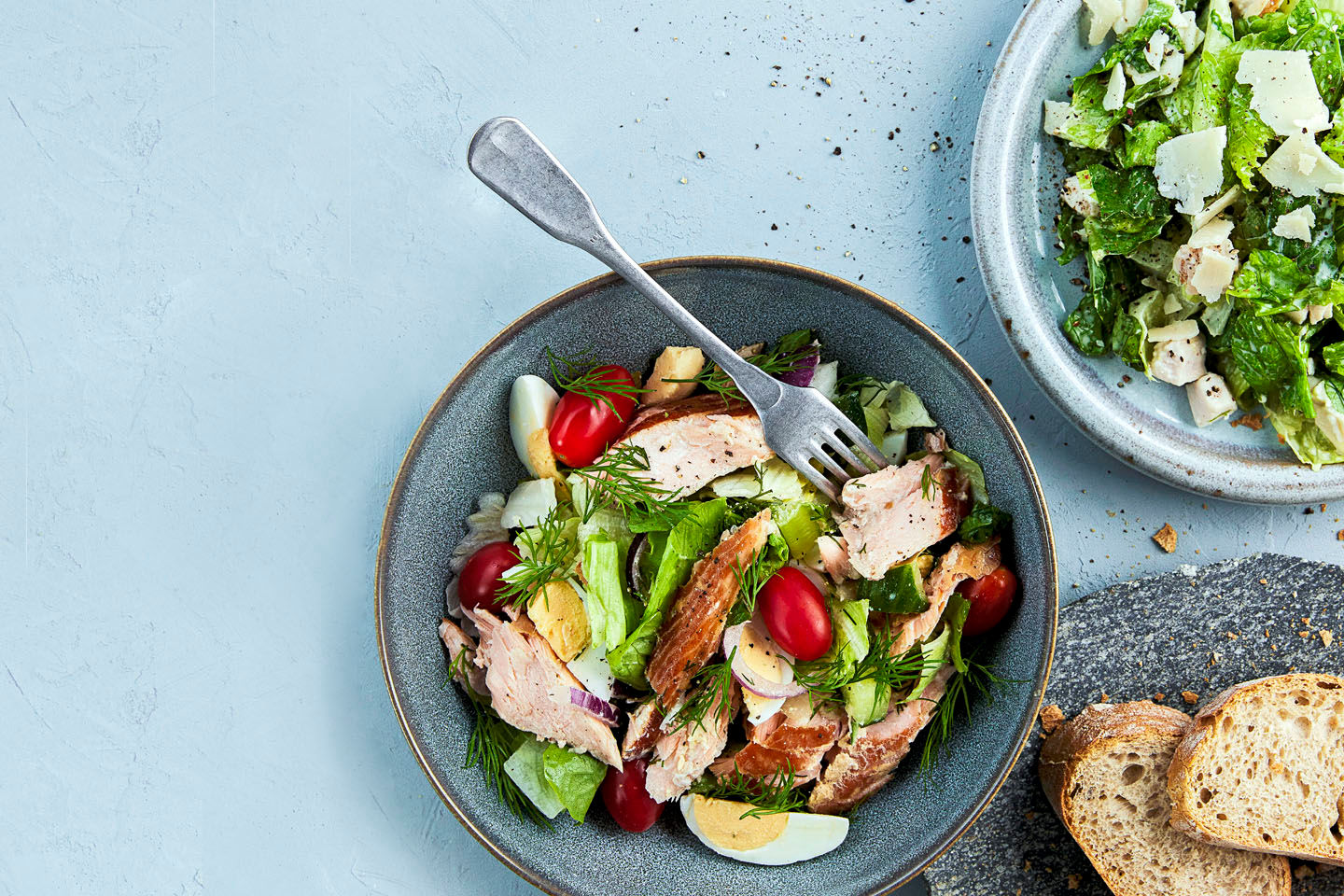 Raikasta salaattia harmaalla lautasella: lohta, kananmunaa, tomaatteja ja vihreää salaattia, vieressä leipää ja toinen salaattilautanen.