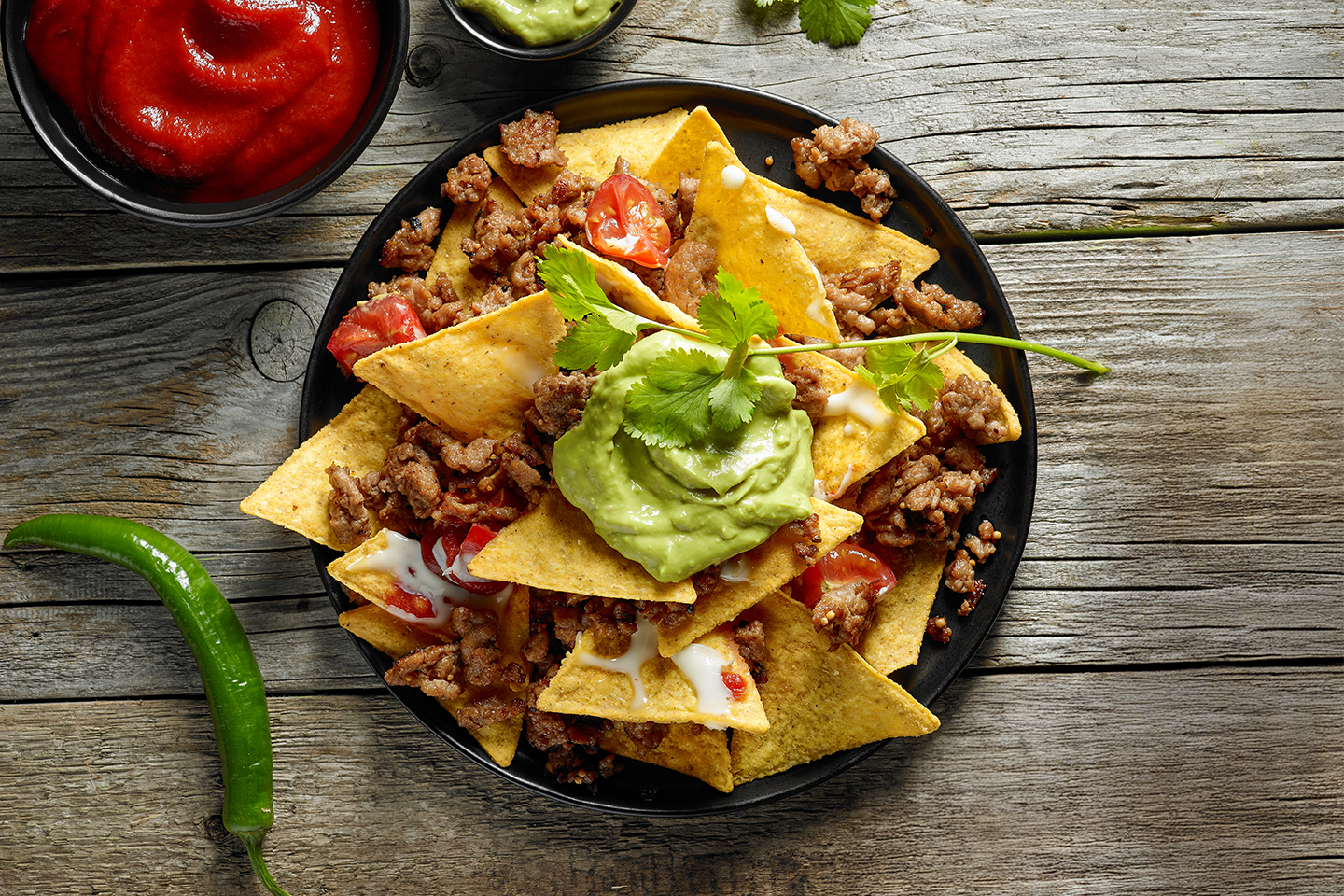 Herkullinen nacholautanen täytetty jauhelihalla, juustolla ja guacamolella, vieressä salsakastiketta ja tuore chili puupöydällä.