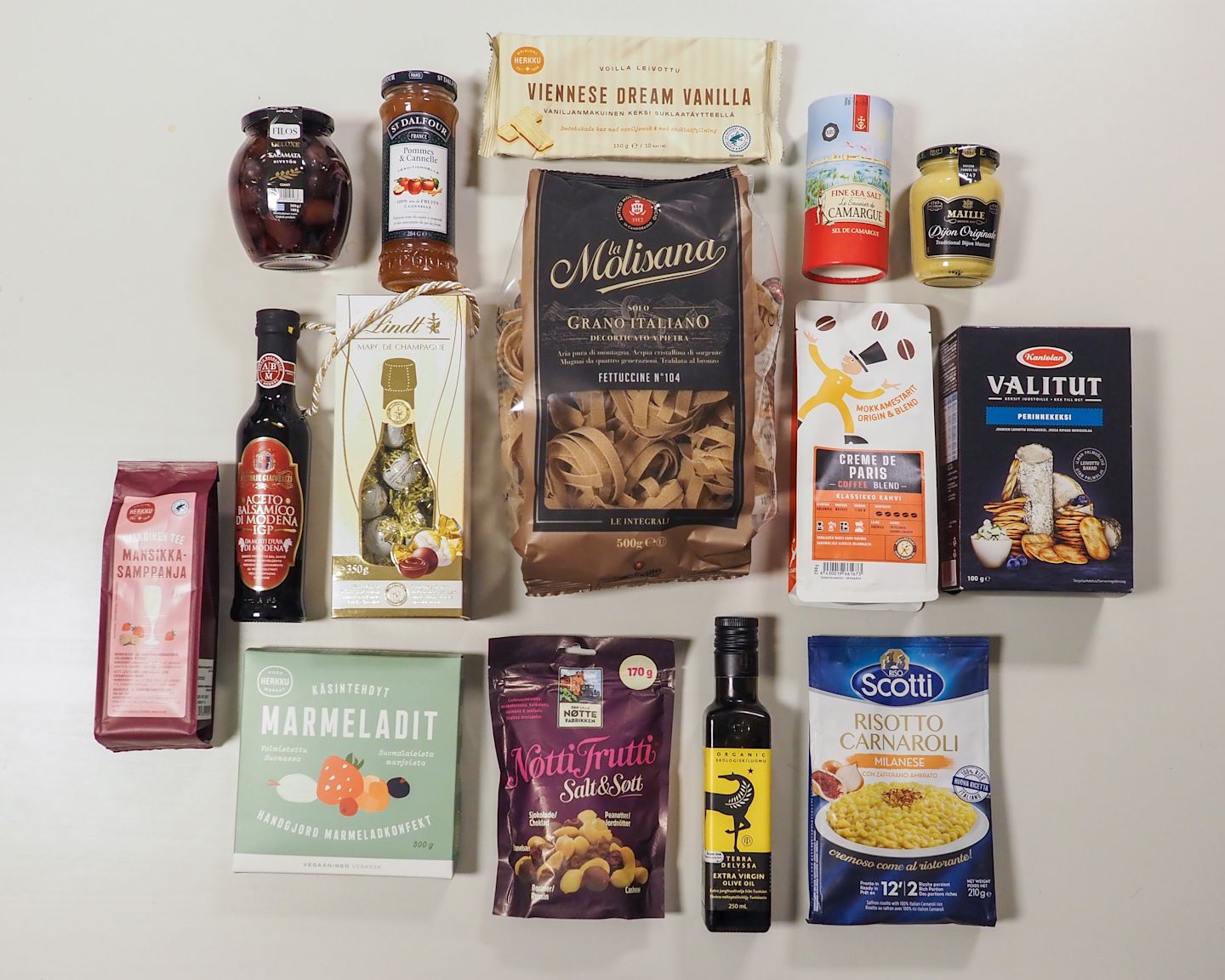 Laadukas valikoima herkkutuotteita, kuten pastaa, suklaata, kahvia, hilloja ja öljyä siististi aseteltuna valkoisella taustalla.