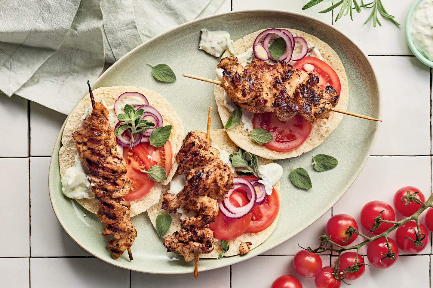 Kolme grillattua kanavarrastaa pitaleivällä, tomaatilla, punasipulilla ja tzatzikilla vaalealla lautasella.