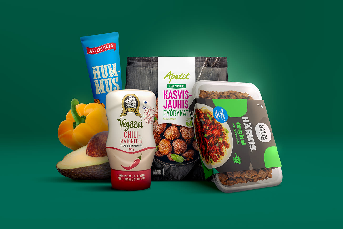 Vihreää taustaa vasten on aseteltu vegaanisia tuotteita: hummusta, chilimajoneesia, kasvisjauhispyöryköitä, härkistä, tuore paprika ja avokado.