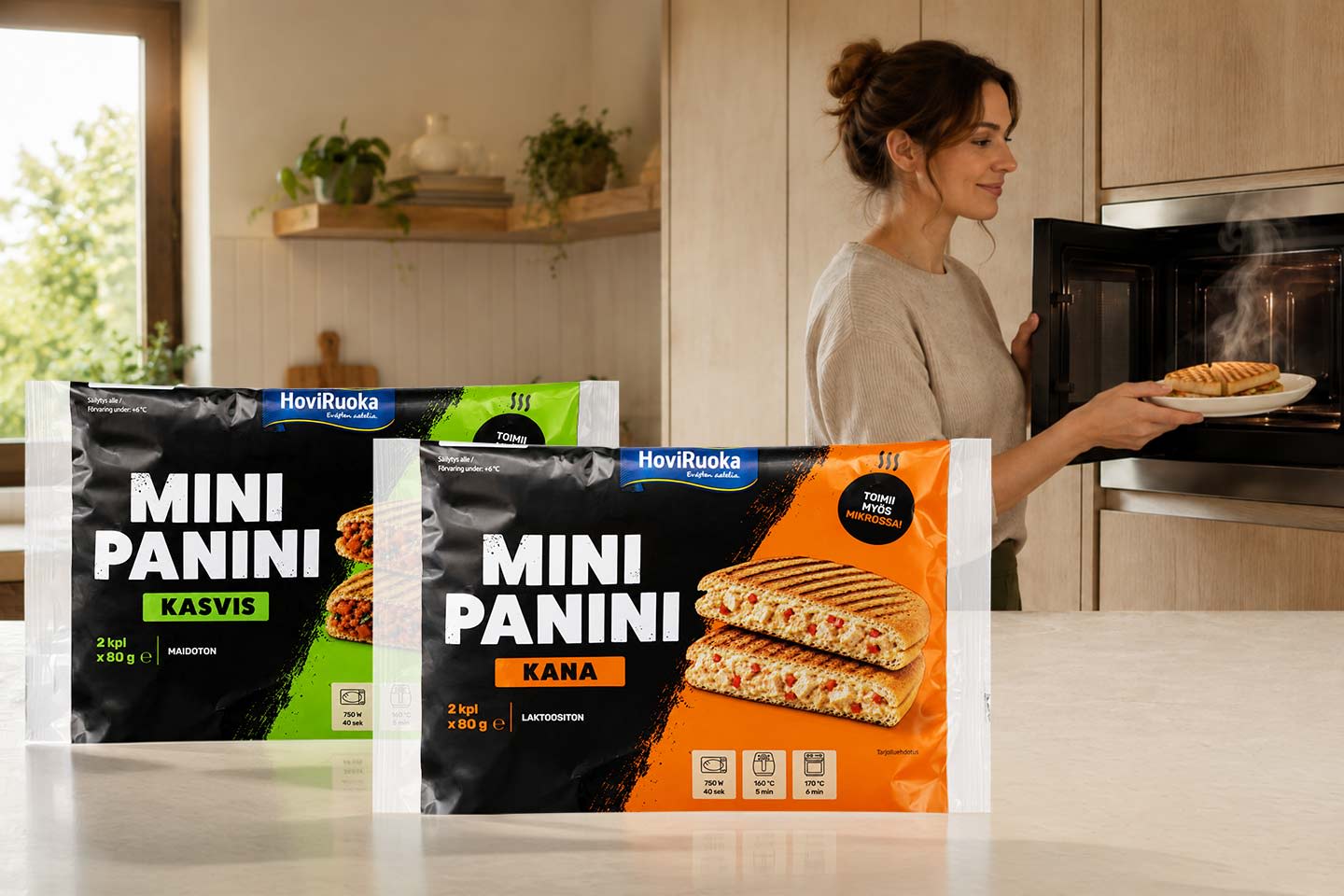 HoviRuoka Mini Panini -kasvis- ja kanapakkaukset keittiön pöydällä, taustalla nainen ottaa höyryävää paninia mikroaaltouunista.