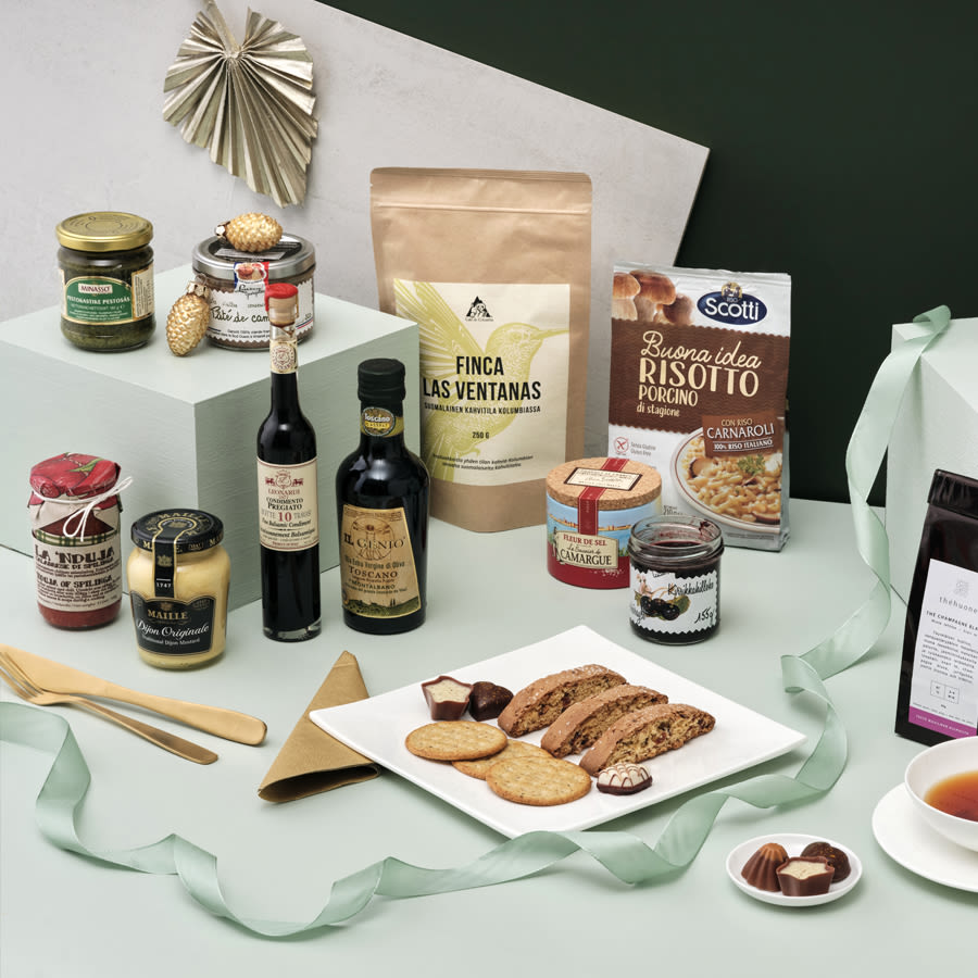 Lahjakoriin sopivia herkkuja aseteltuna vaalealle pinnalle: keksejä, risottoriisiä, oliiviöljyä, balsamicoa, hilloja, levitteitä, suolaisia ja makeita naposteltavia sekä suklaata, kaikki koristeltuna juhlanauhoin.