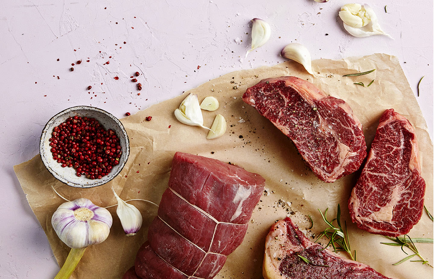 Lihaa leivinpaperin päällä. Ympärillä pippuria kulhossa, valkosipulia ja yrttejä.