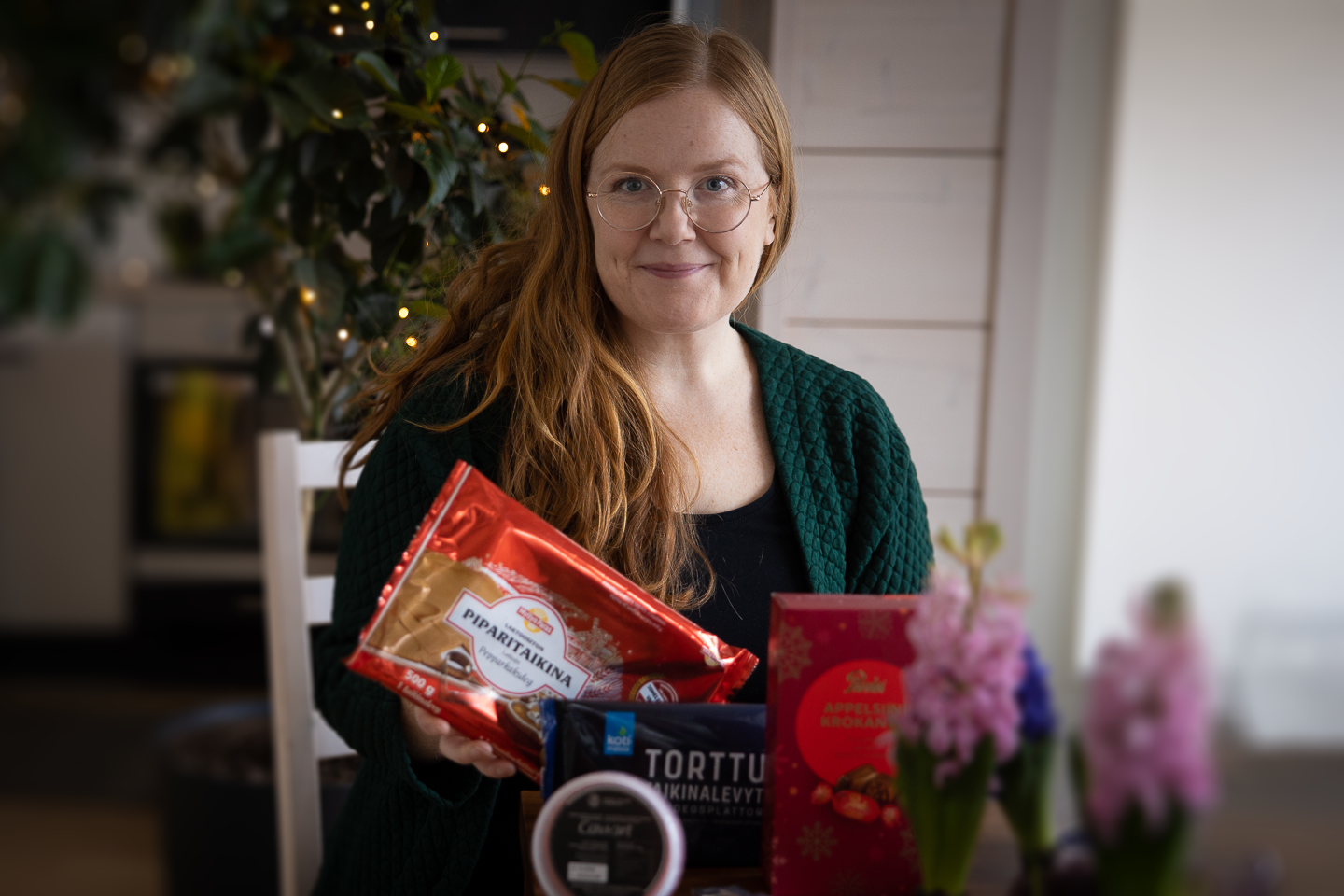 Hymyilevä henkilö vihreässä neuletakissa pitelee leipomistarvikkeita, taustalla näkyy viherkasvi ja jouluvaloja.