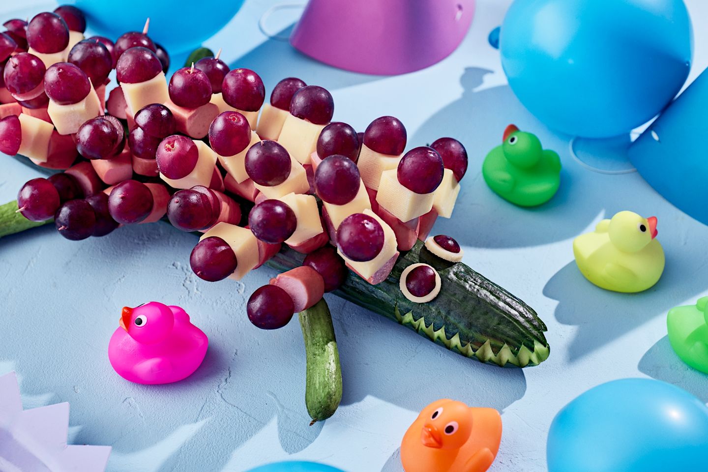 Krokotiilin muotoinen ruoka-asettelu, jossa viinirypäleitä , nakkeja ja juustoa tikuilla kurkun päällä, ympärillä värikkäitä muoviankkoja.