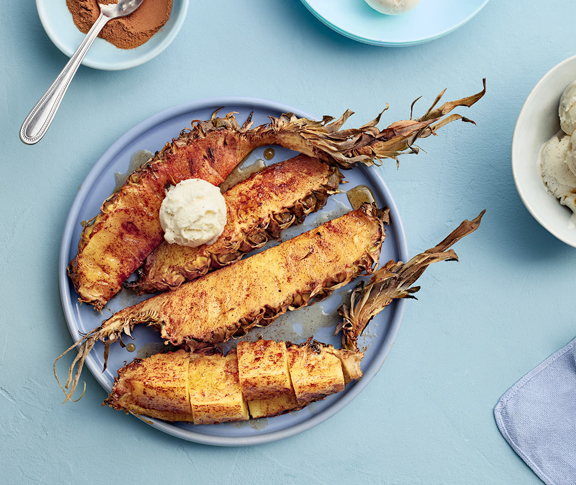 Grillattua ananasta lohkoina sinisellä lautasella. Päällä jäätelöpallo ja vieressä kanelia kulhossa.