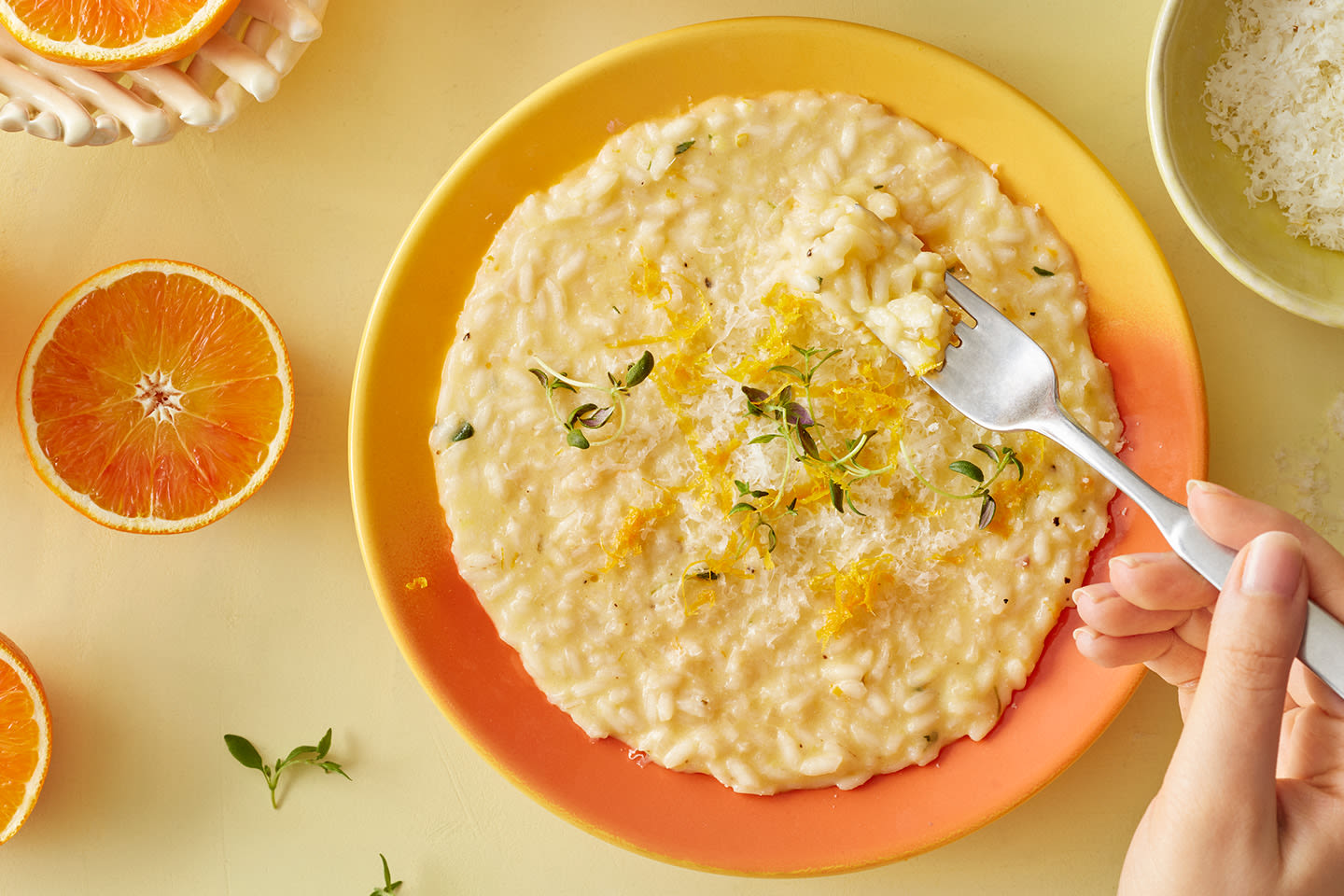 Kermaisella keltaisella lautasella sitruunarisottoa, jota koristavat yrttiversot ja appelsiiniraaste, vieressä tuoreita appelsiinilohkoja.