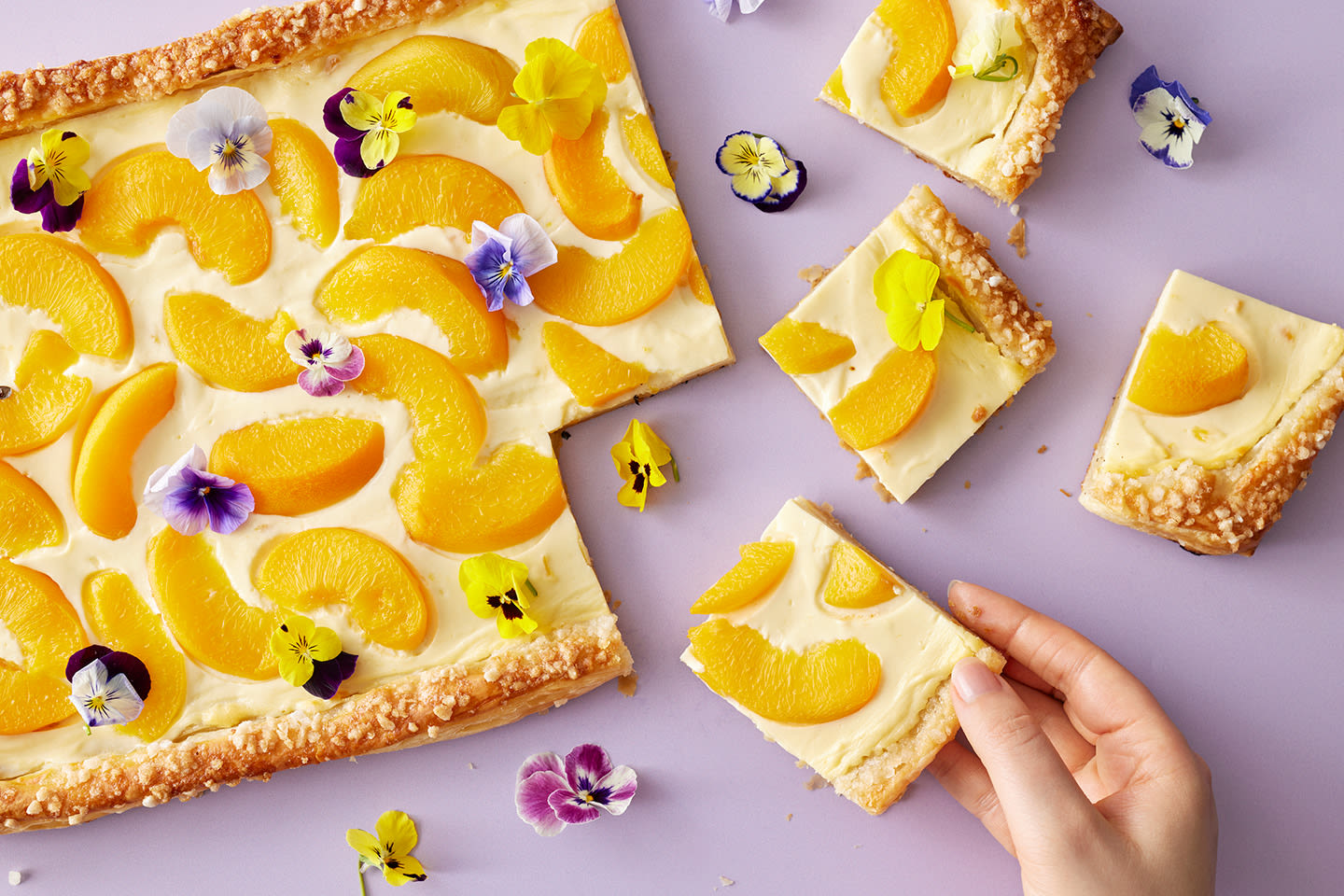 Persikkapiirakka koristeltuna tuoreilla orvokinkukilla ja persikanviipaleilla, käsi ottaa palan piirakkaa.