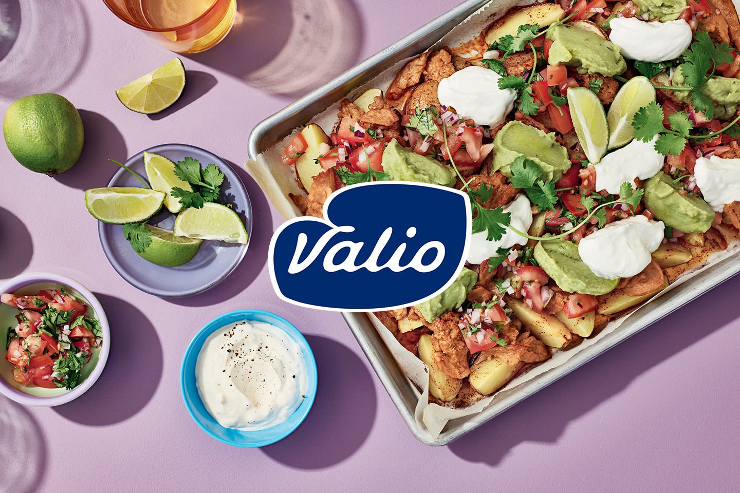 Meksikolaistyyliset nacholautanen täynnä kanaa, avokadoa ja kermaa, vieressä limeä ja salsaa, keskellä Valio-logo.