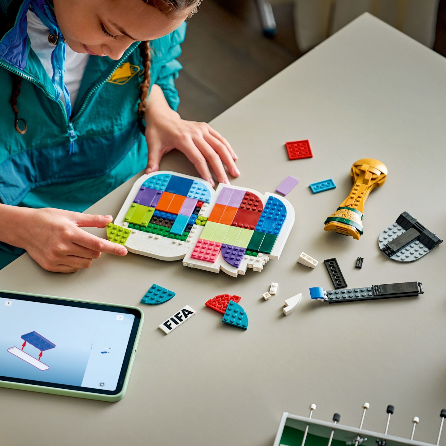 Lapsi rakentaa värikkäästä LEGO-palasista FIFA-aiheista settiä pöydällä, vieressä tabletti ja kultainen pokaali.