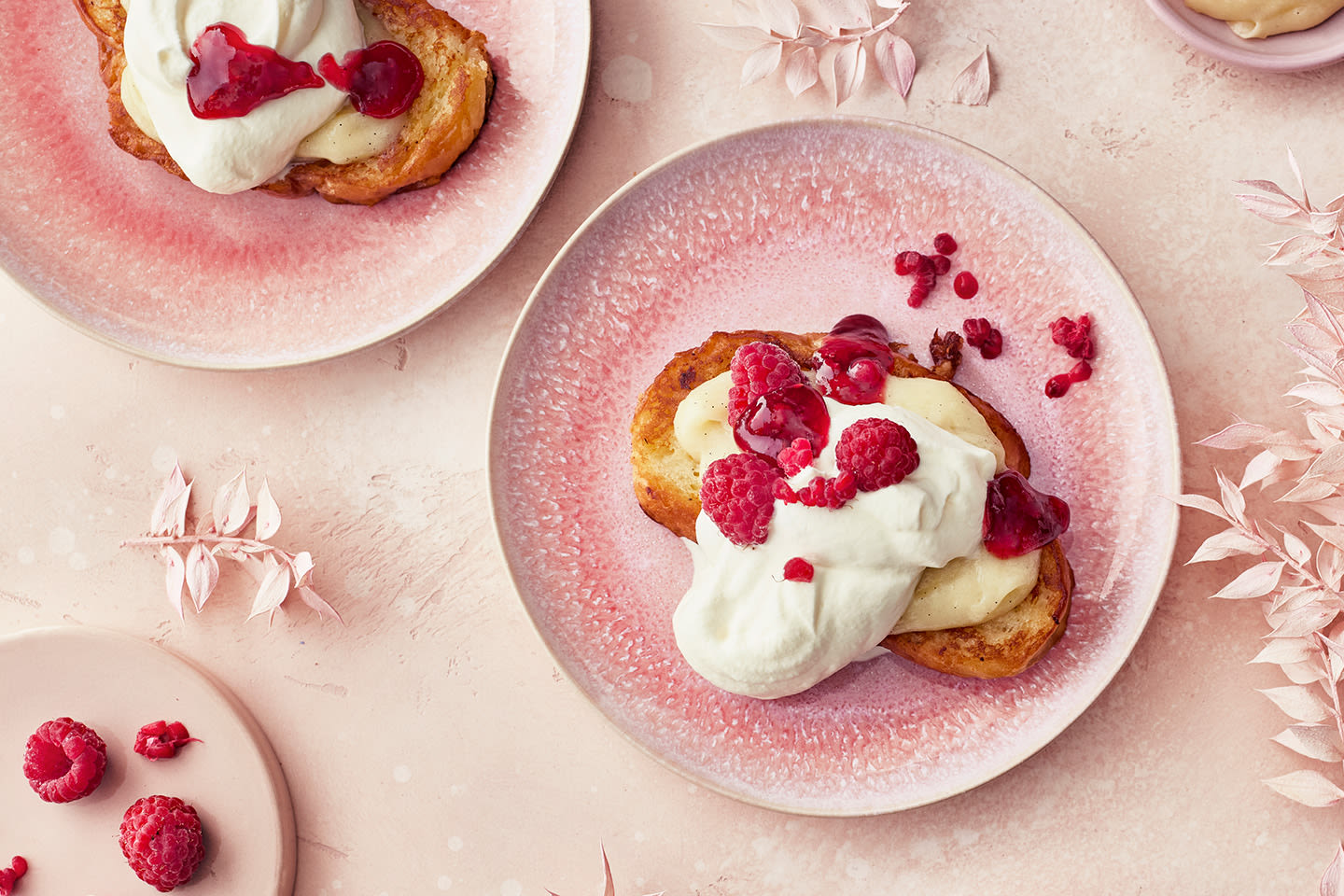 Herkullisia ranskalaisia leipiä vaaleanpunaisilla lautasilla, päällä kermaa, banaania ja tuoreita vadelmia.