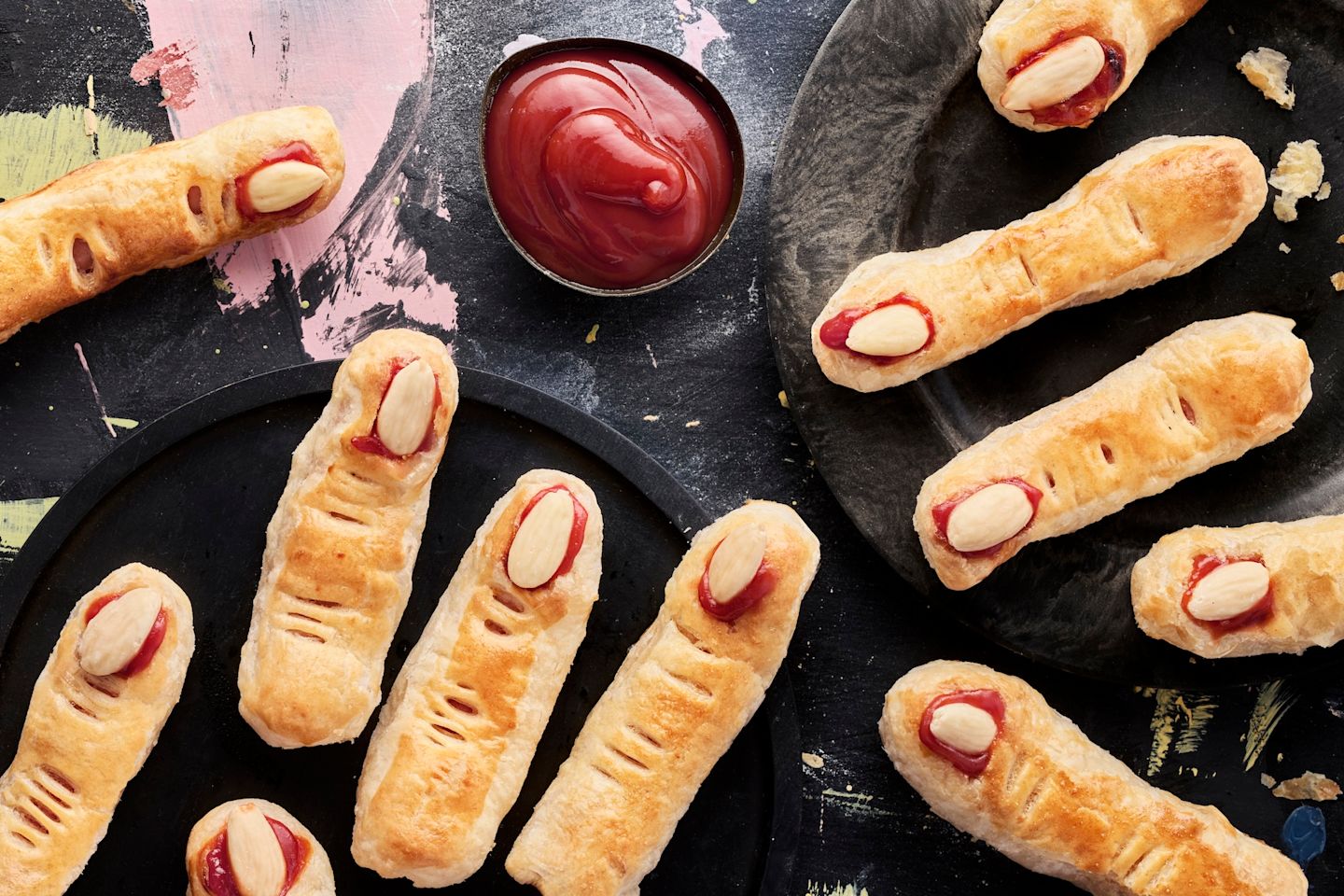 Halloween-leivokset, jotka näyttävät sormilta – paistettua lehtitaikinaa pitkulaisessa muodossa, kynsinä mantelit ja vieressä ketsuppia pienessä dippiastiassa.