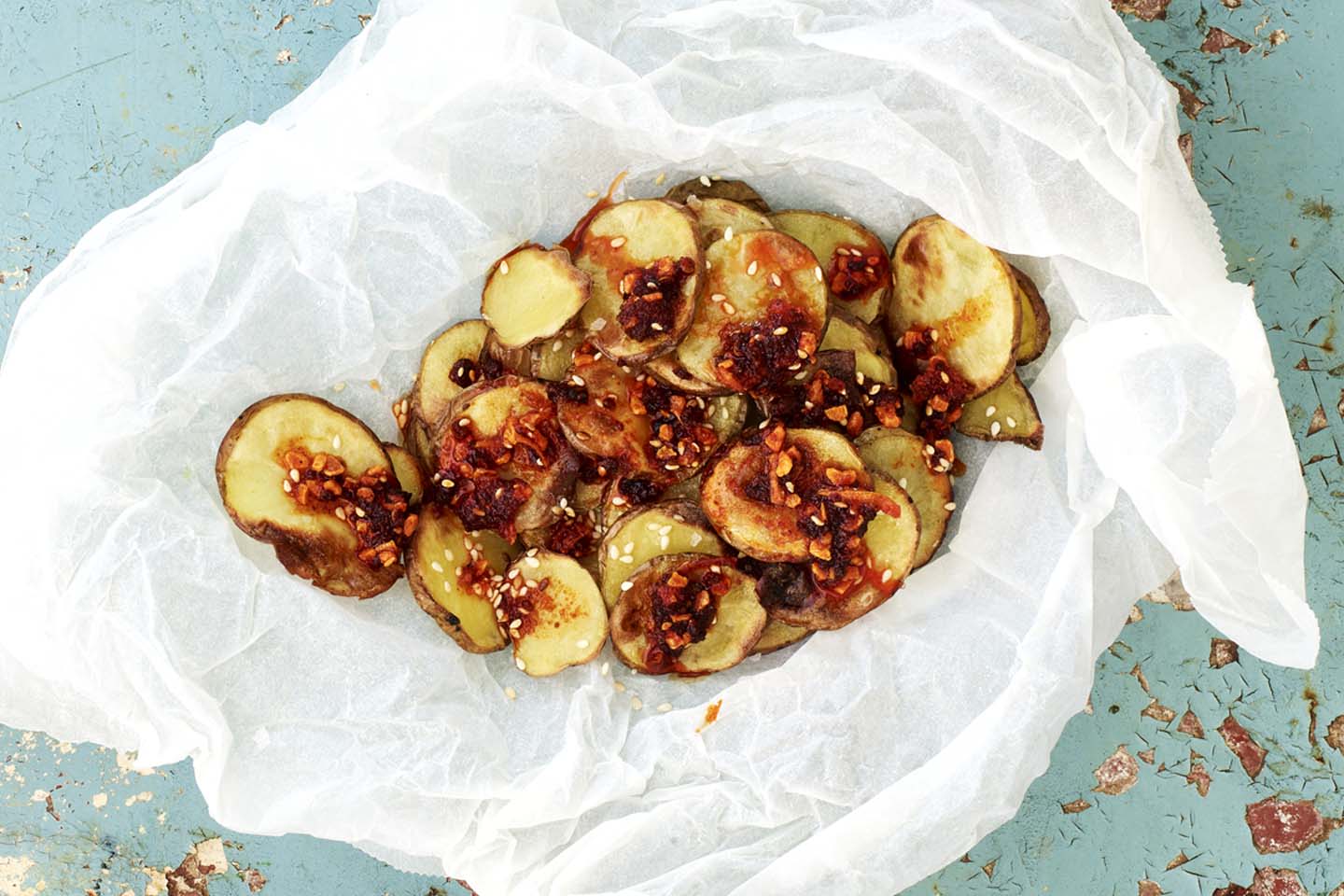 Paistettuja perunaviipaleita leivinpaperilla sinertävällä pinnalla, päällä chili- ja valkosipuliöljyä ja seesaminsiemeniä.