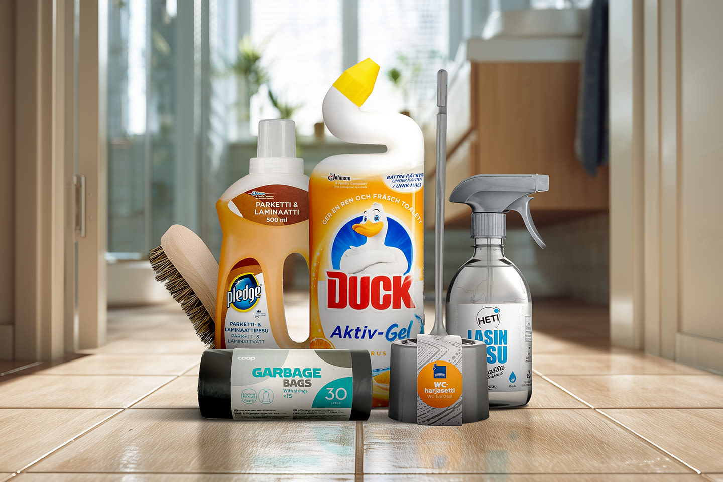 Kodin siivoustarvikkeita lattialla: puhdistusaineita, harja, roskapusseja ja WC-paperia valoisassa tilassa.