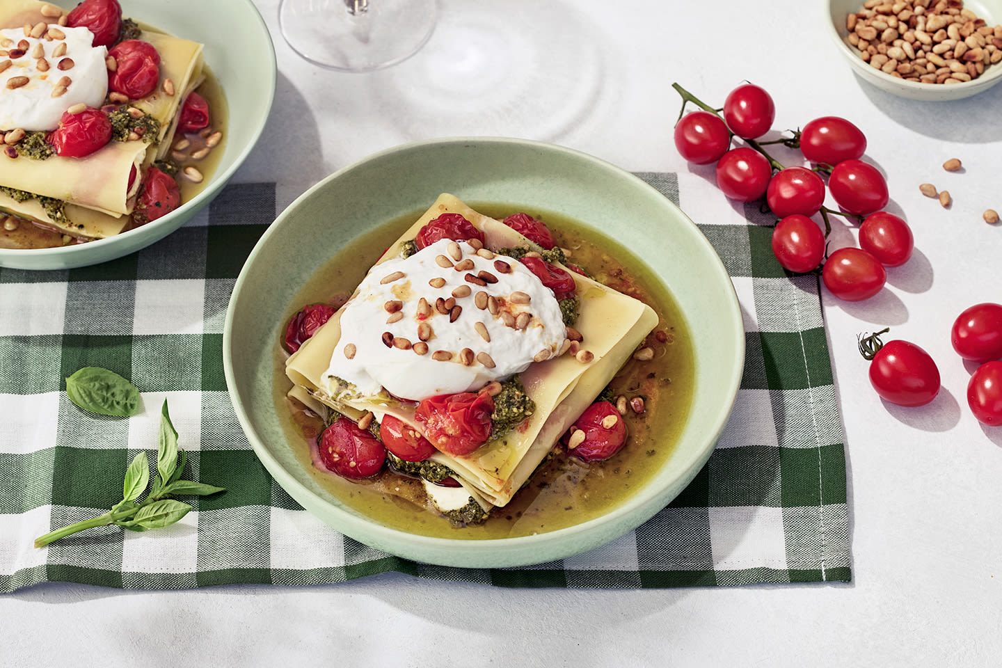 Vihreässä kulhossa lasagnea pestolla, kirsikkatomaateilla, burratalla ja pinjansiemenillä, taustalla tuoreita tomaatteja.