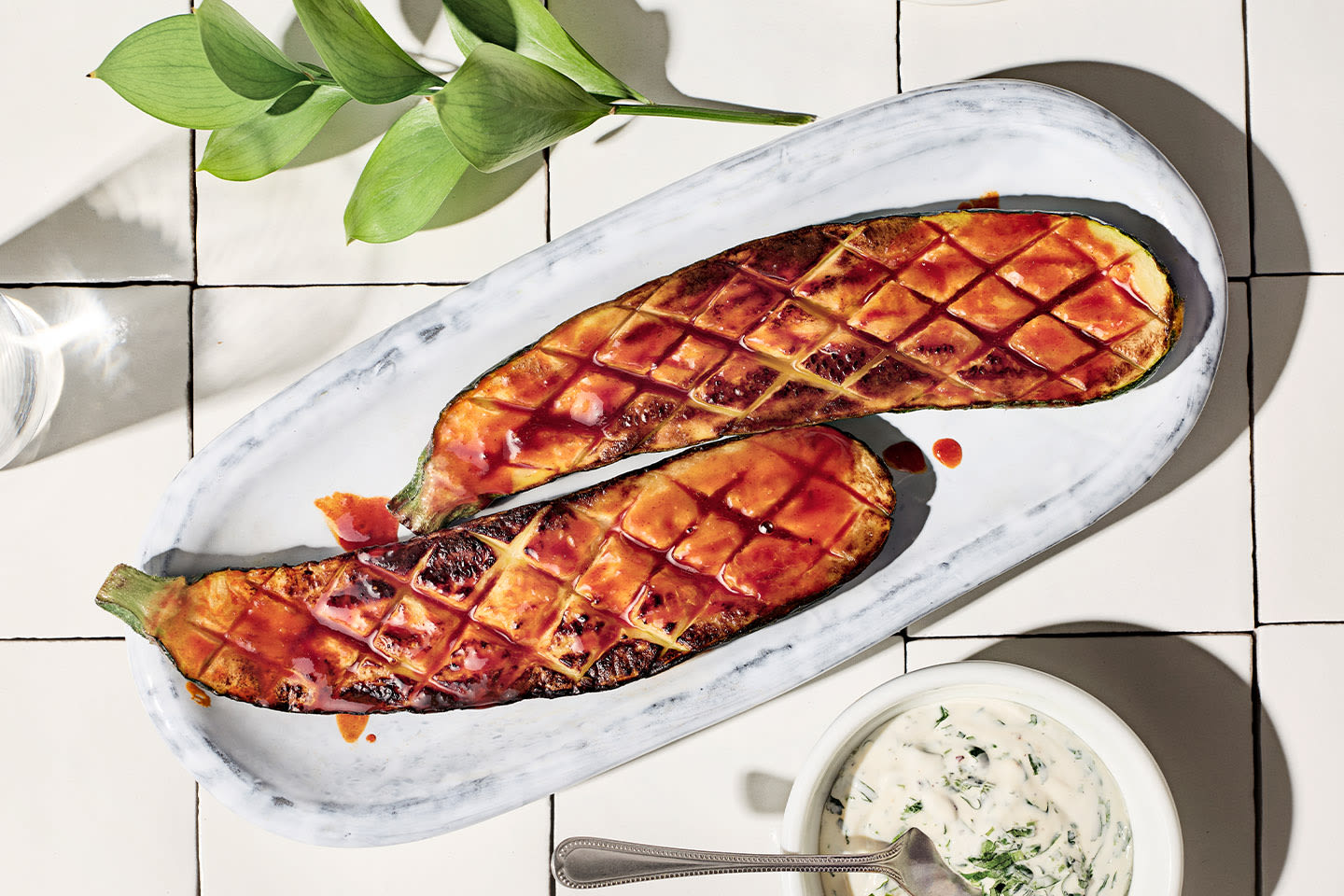 Kaksi grillattua kesäkurpitsaa puoliksi leikattuna marmorivadilla, kastettuna punaisella kastikkeella, vieressä yrttijogurttikastike.