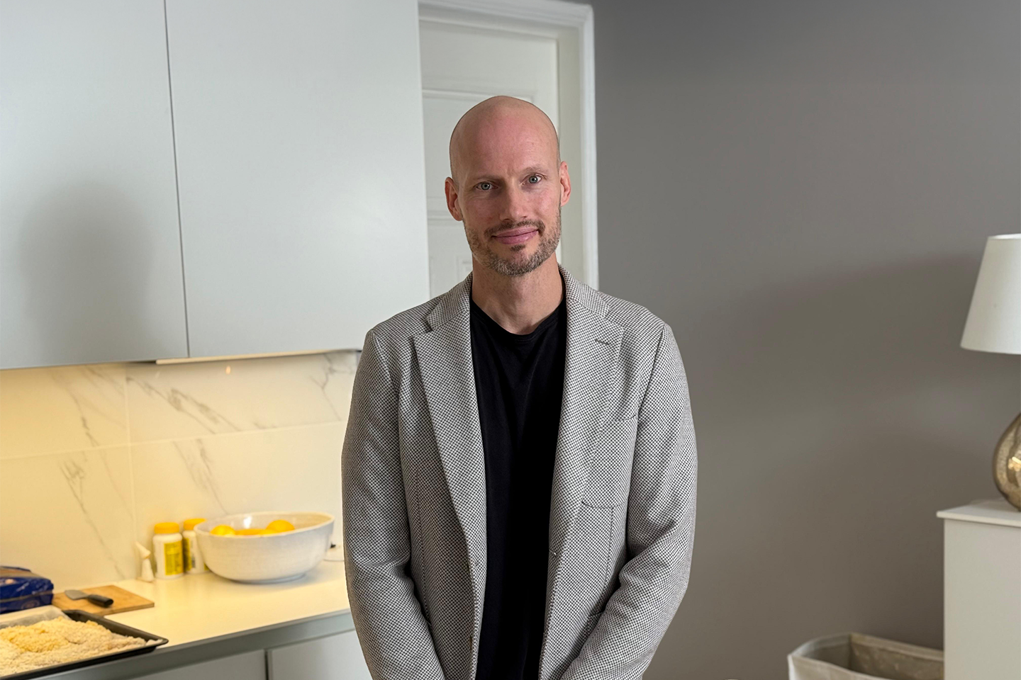 Henkilö harmaassa takissa ja mustassa paidassa seisoo modernissa keittiössä, jossa on marmoripintoja ja kulhollinen keltaisia hedelmiä.