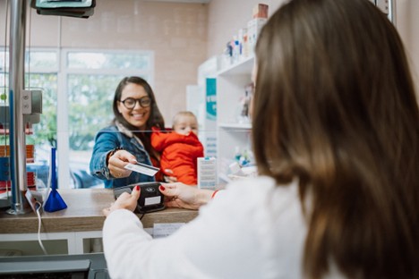 A woman holding a baby uses her card to pay for a prescription from a pharmacist